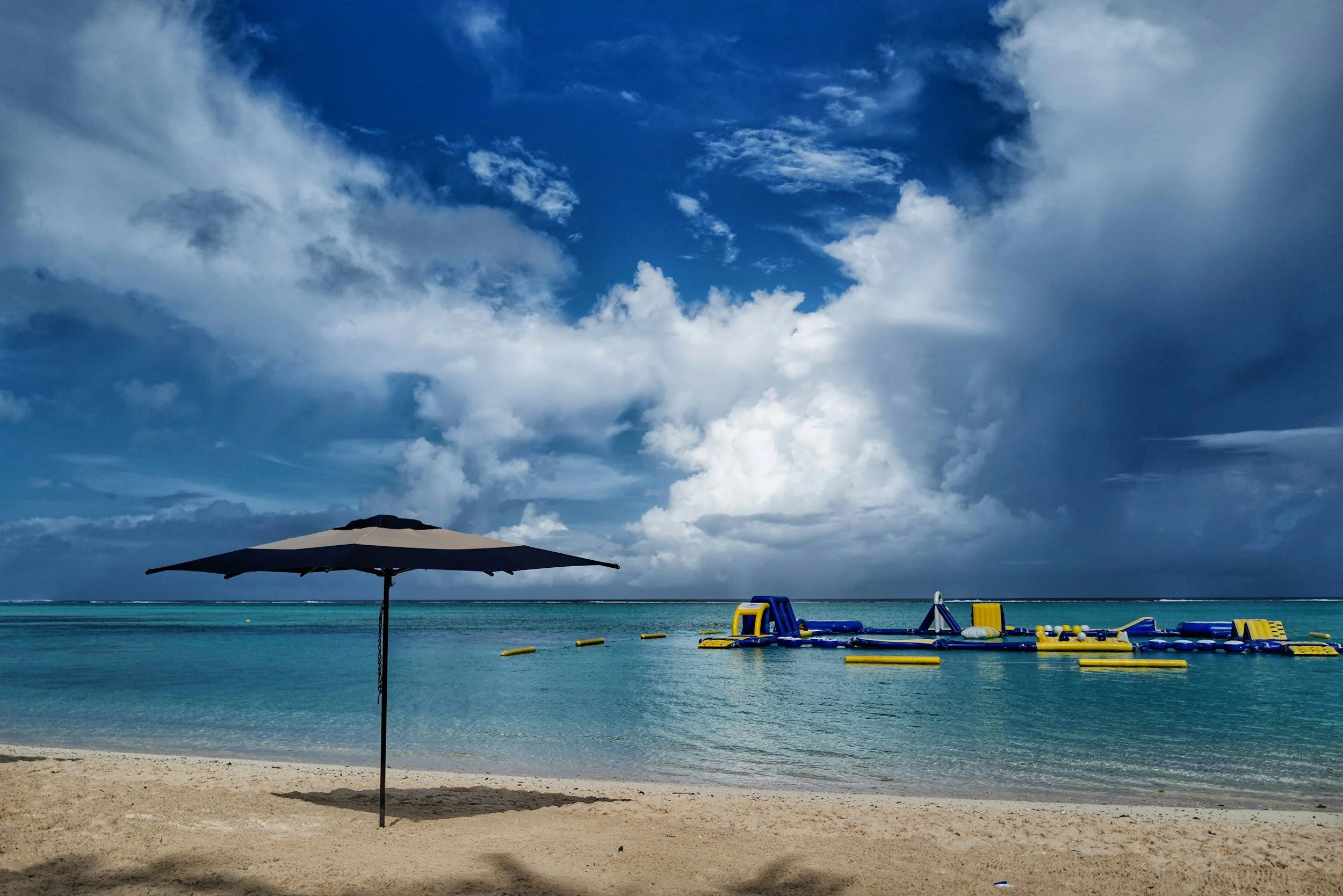 Beach scene with a large umbrella on sandy shore, floating floating water toys, calm ocean, and a sky with scattered clouds.