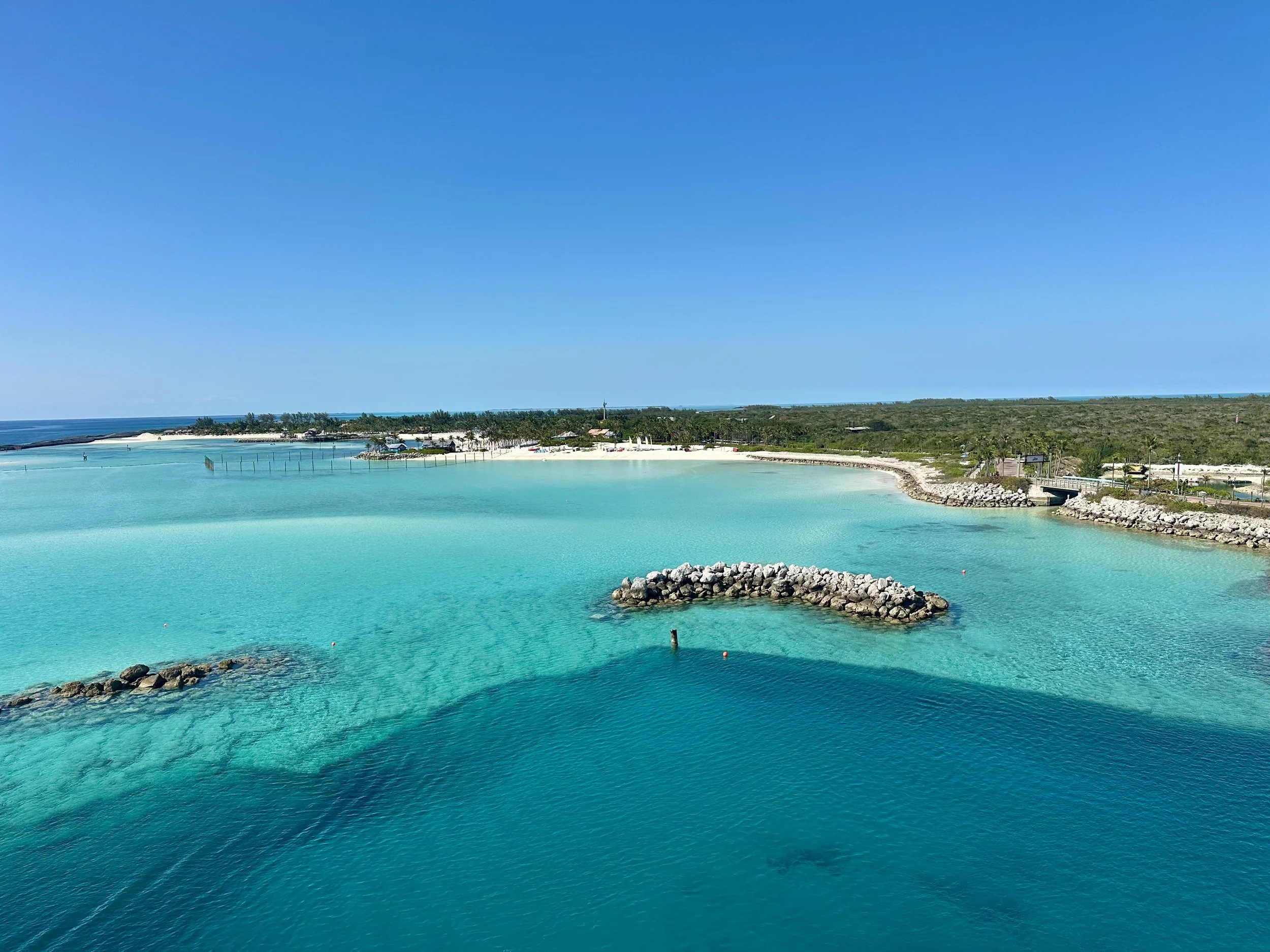 Clear turquoise ocean water with a small rocky jetty, a sandy beach, and a distant shoreline with trees and buildings under a bright blue sky.