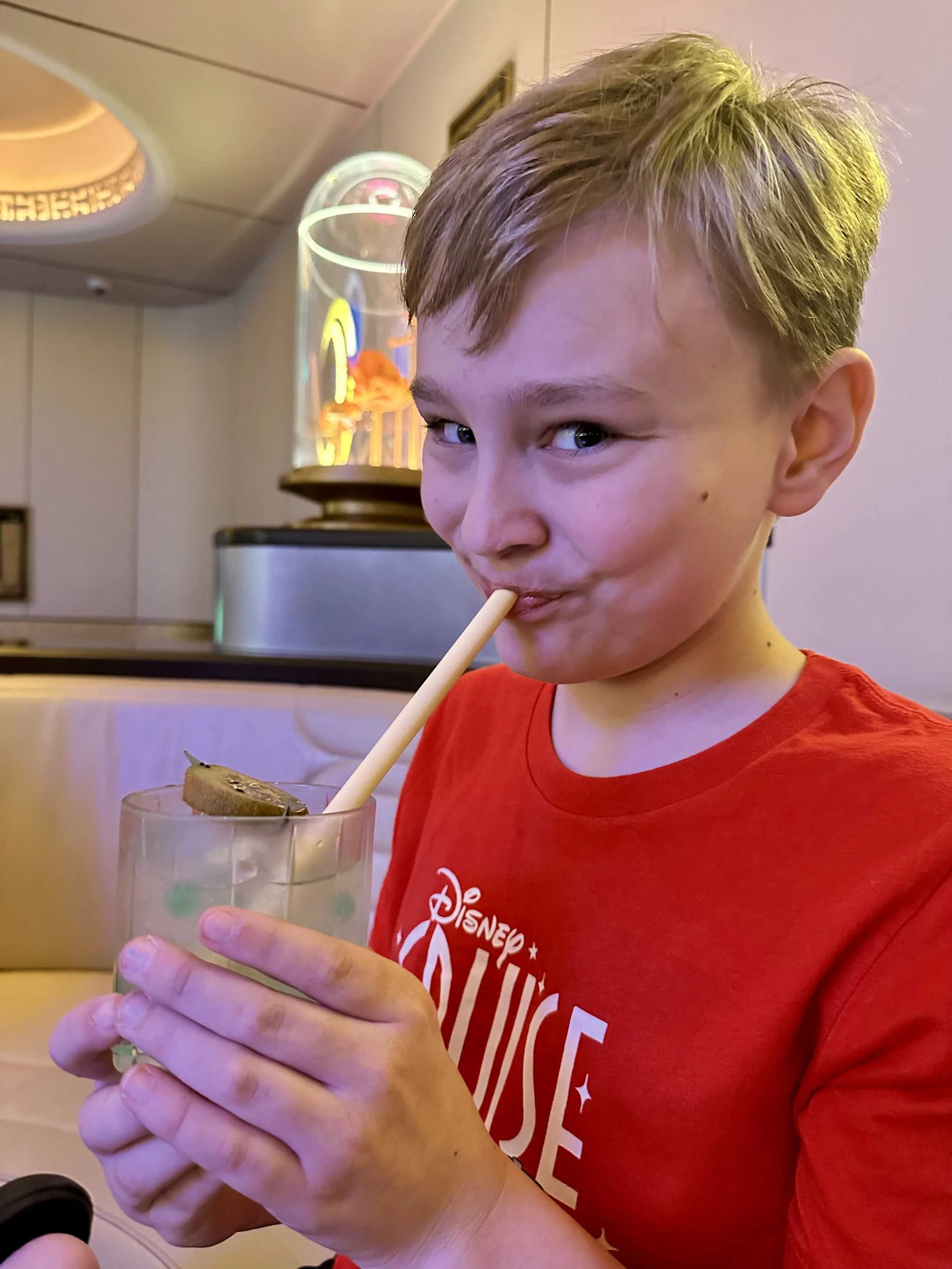 A young boy in a red Disney shirt is holding a glass of a green beverage with a straw and a cracker with spread on it, sipping through the straw.