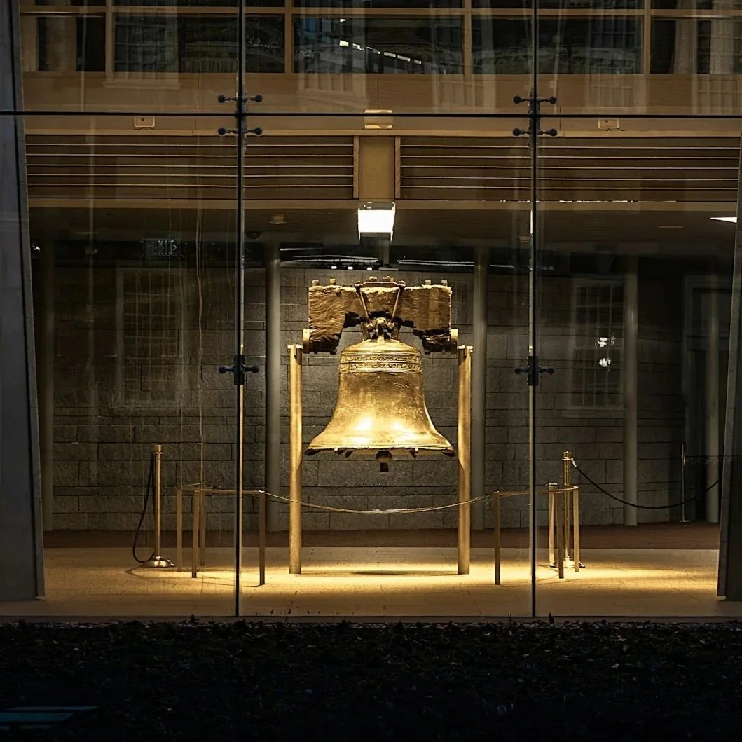A large bronze bell displayed inside a glass case with a metal frame, roped off with stanchions, in a museum or gallery setting.