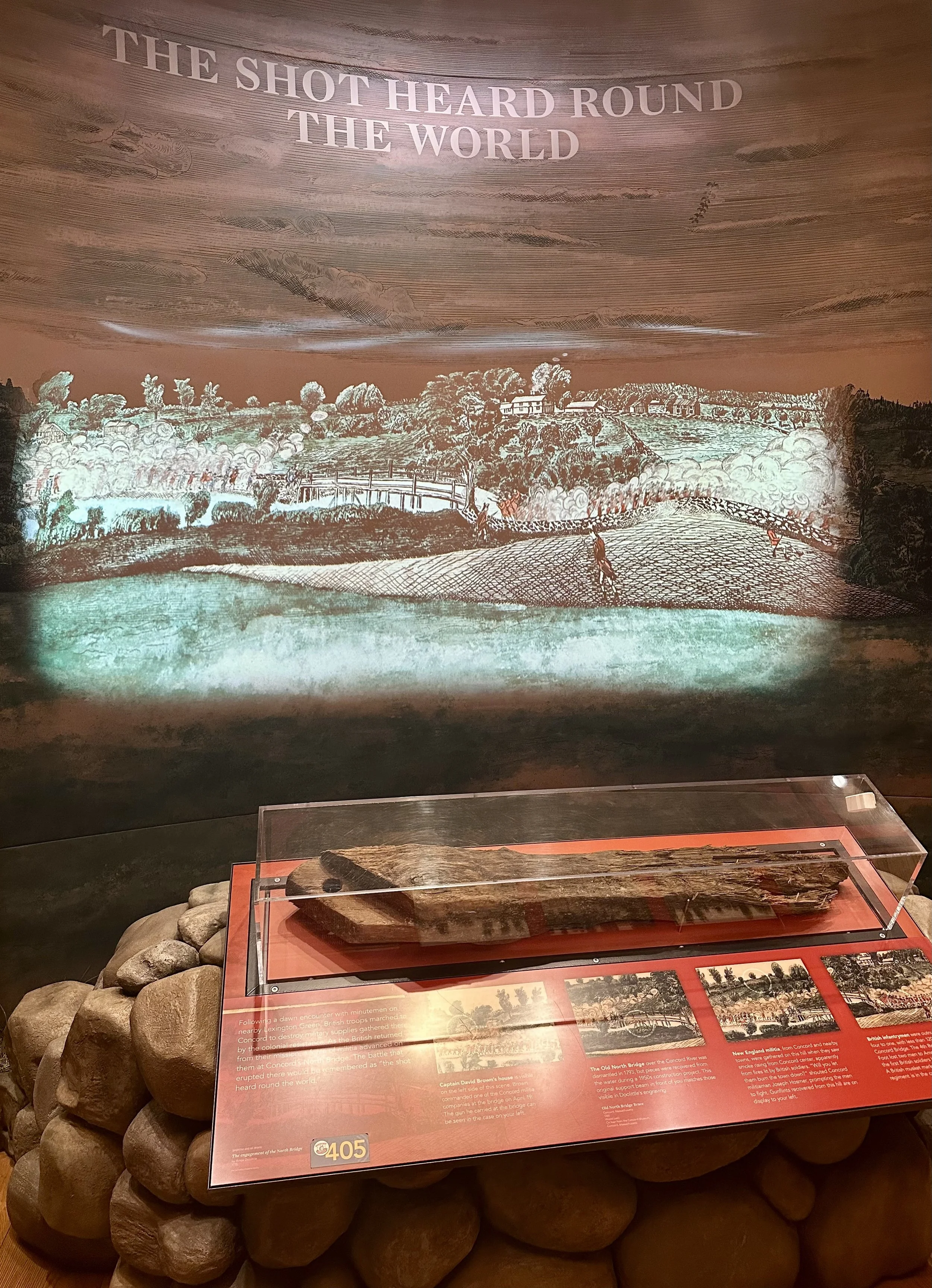Historical exhibit featuring an illustration of a battlefield scene with soldiers and a caption about the engagement of the North Bridge during the American Revolution, with a wooden musket displayed in front of a glass case.