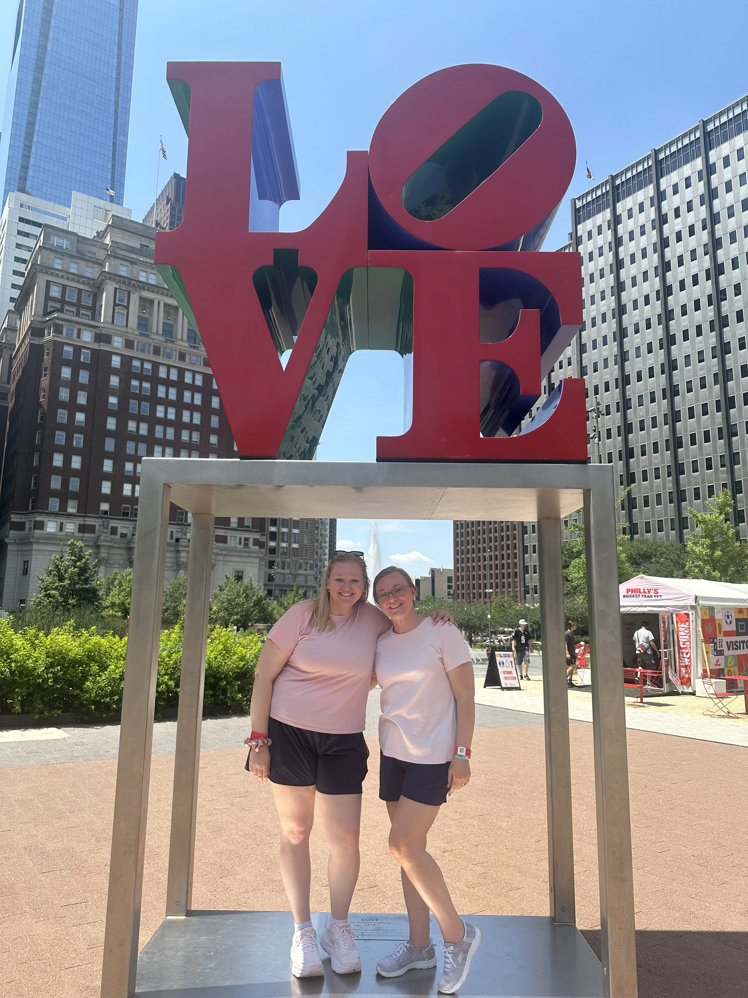 Two smiling women with long hair standing inside a large, red and black sculpture of the word "LOVE" at JFK Plaza park in Philadelphia with tall buildings and clear blue sky in the background.