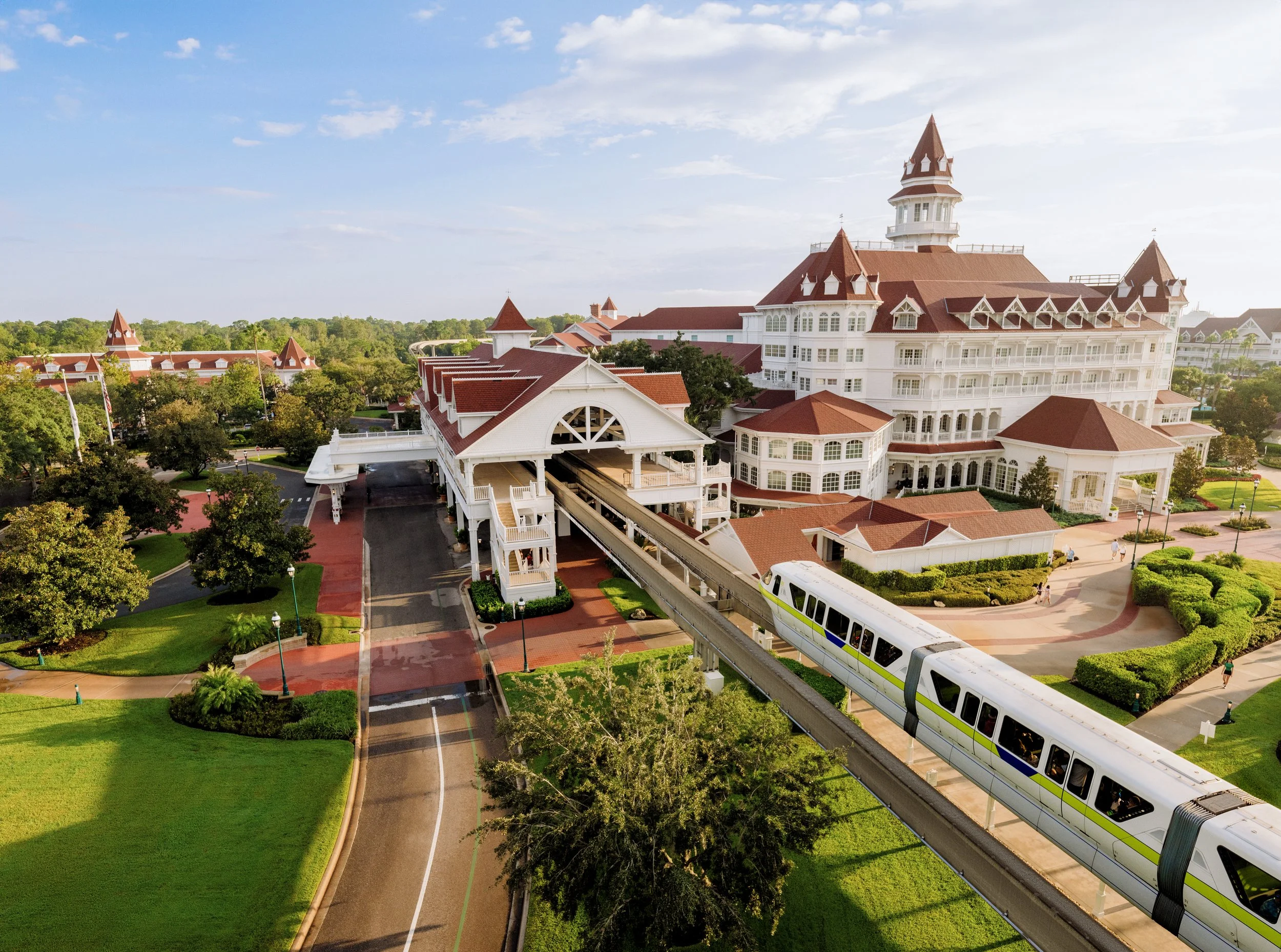 A large, historic hotel with white exterior and red roofs, surrounded by lush green trees and gardens, featuring a monorail train passing on an elevated track nearby.