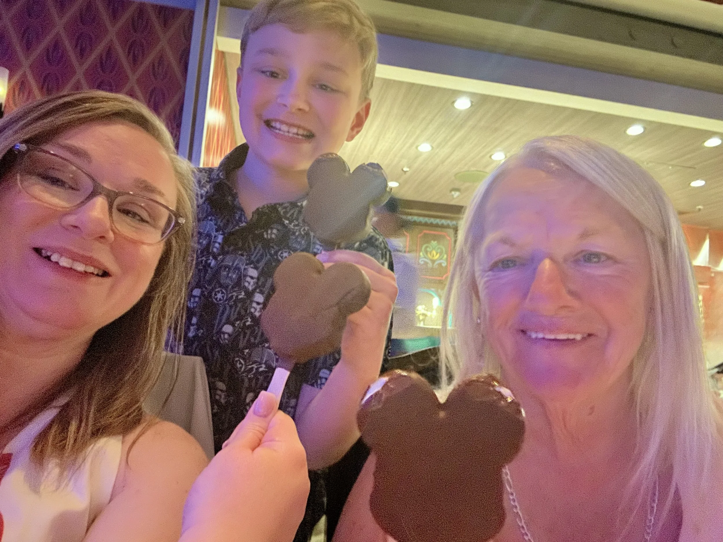 A group of people smiling and taking a selfie together in a brightly lit indoor setting, with a young boy holding a brown heart-shaped object.