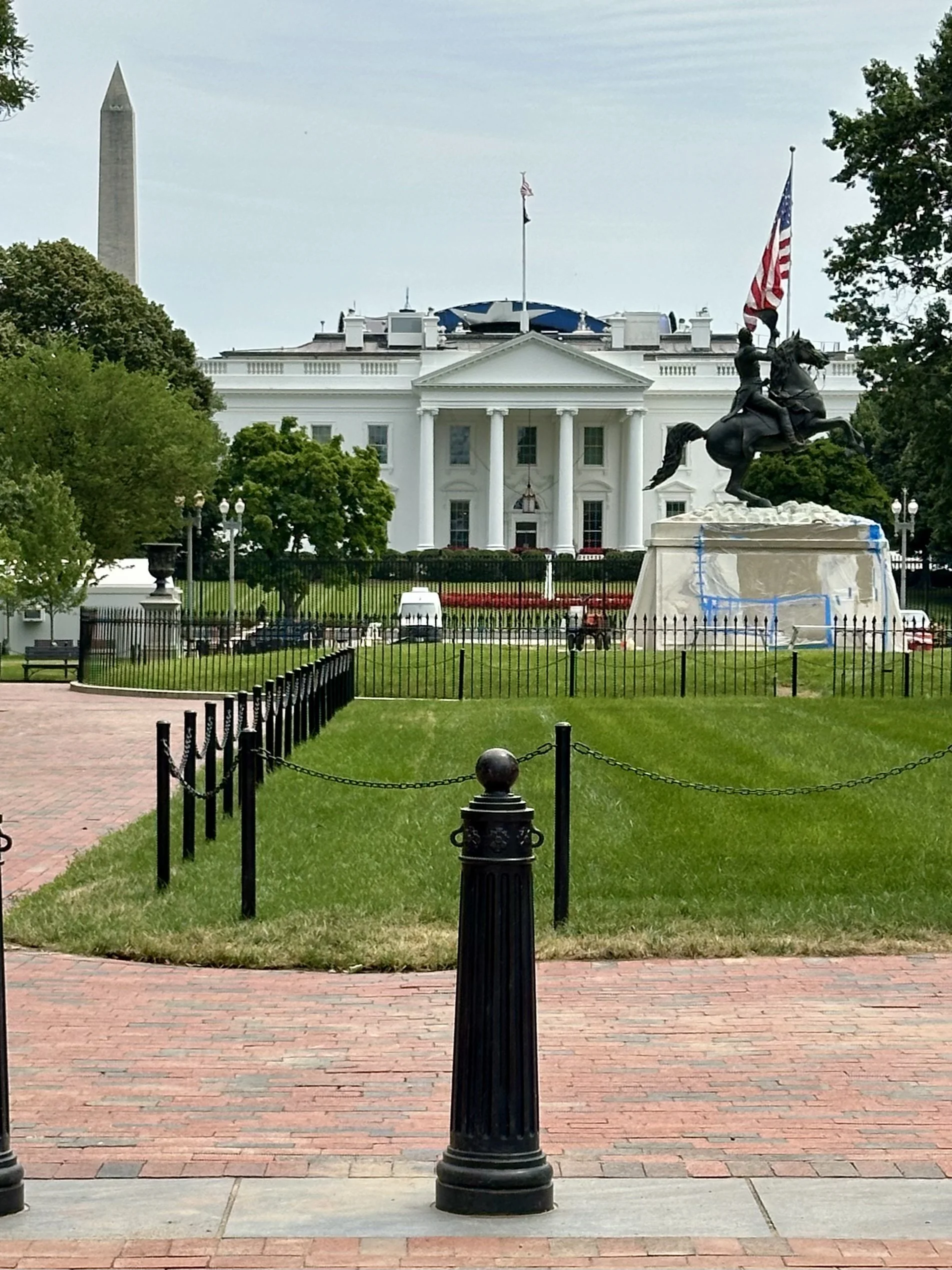 View of the White House with a statue of a man on a horse holding an American flag in front of it. The area is surrounded by trees, and there is a brick walkway and a black fence in the foreground.