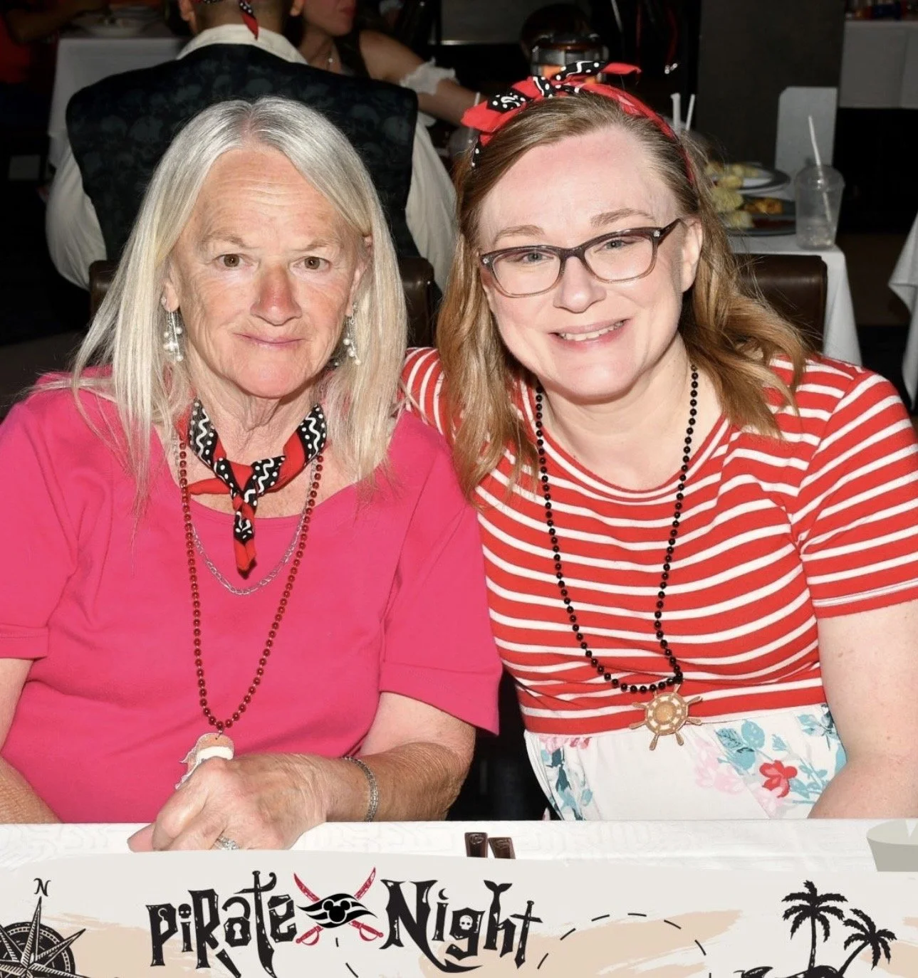 Two women smiling at a party called Pirate Night. One woman has long blonde hair, wearing a pink shirt, with a black and red bandana around her neck and black beaded necklace. The other woman has red hair, glasses, and wears a red and white striped s