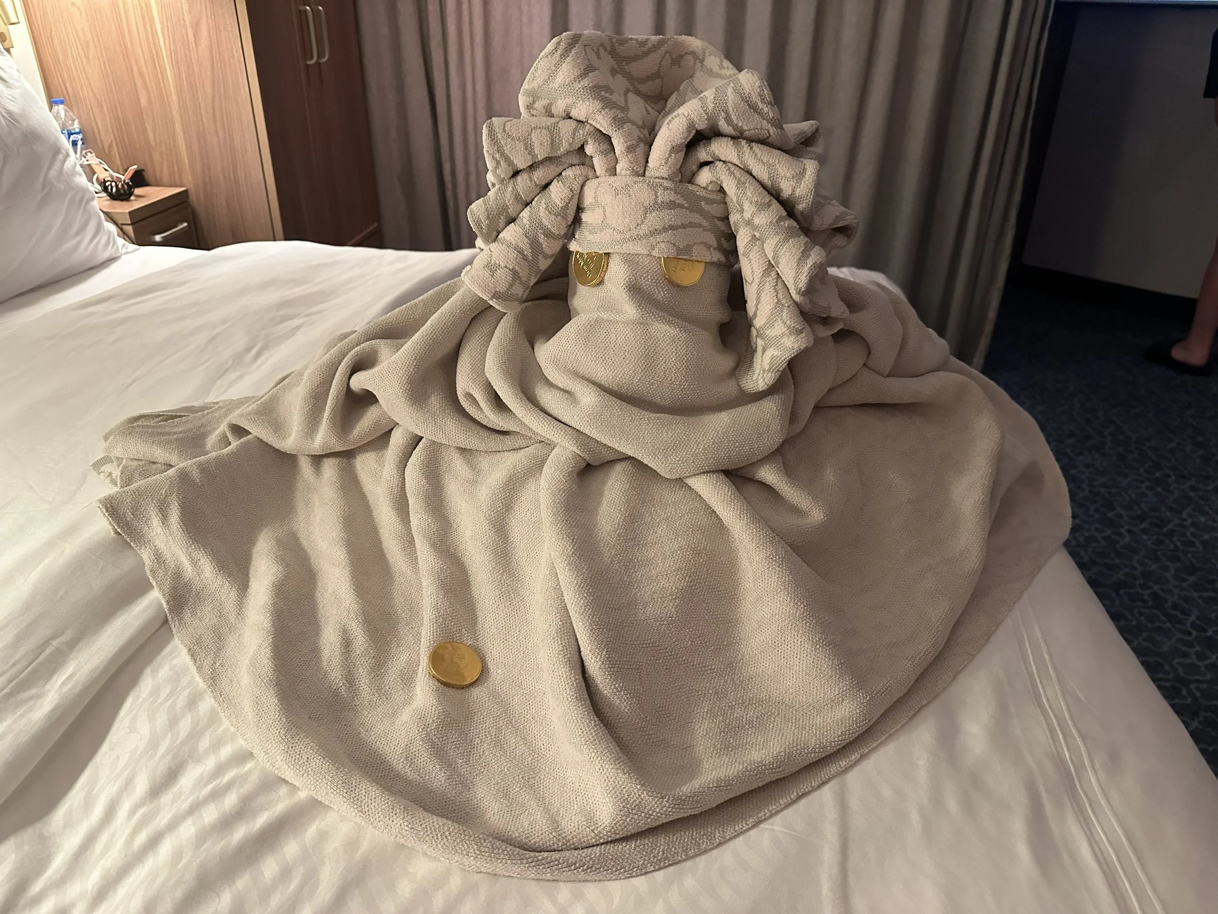 A towel folded to resemble a person, with a bracelet, coins, and ring placed on it, on a bed in a hotel room.