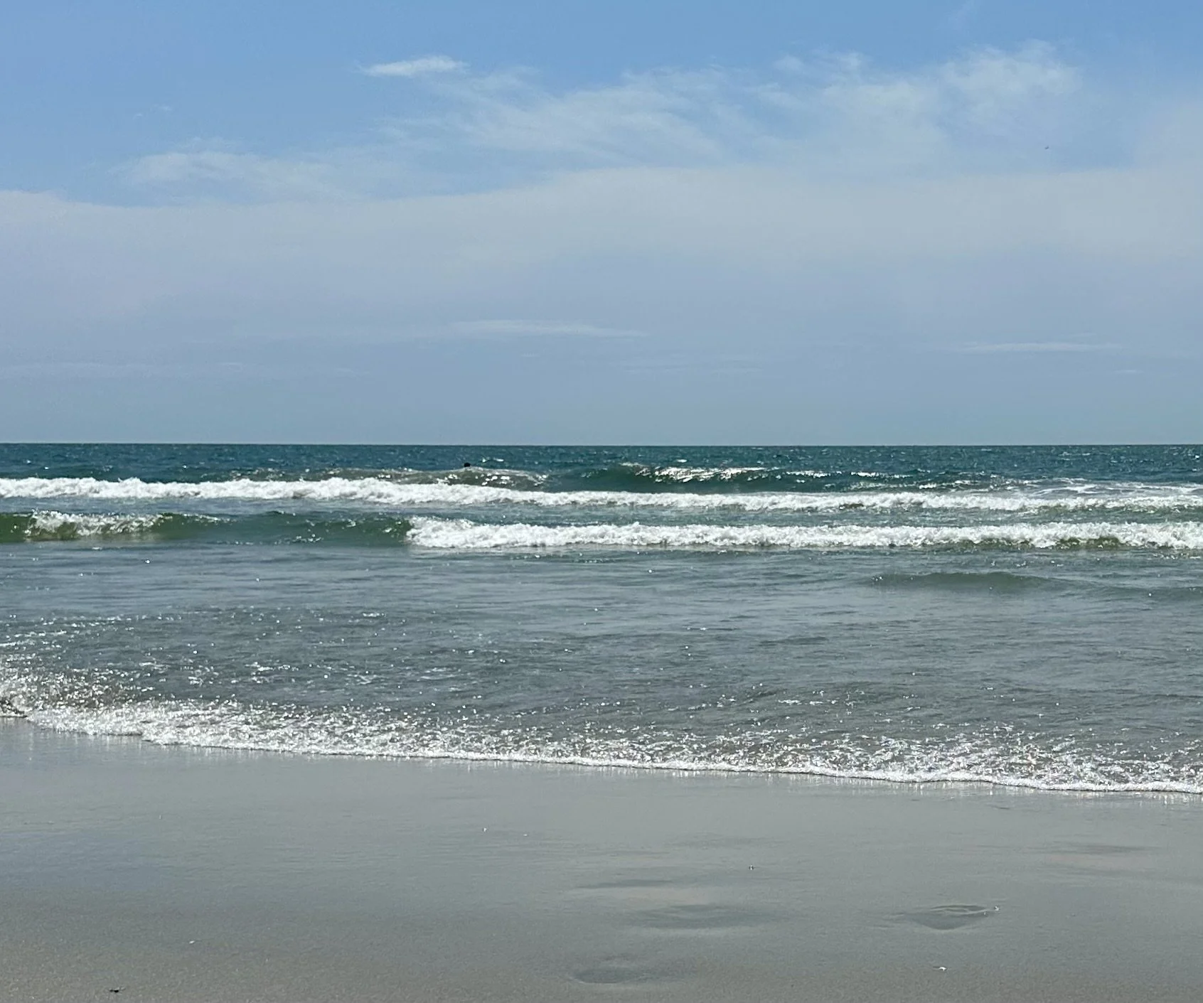 Photo of an ocean with small waves crashing onto a sandy beach under a blue sky with a few clouds.