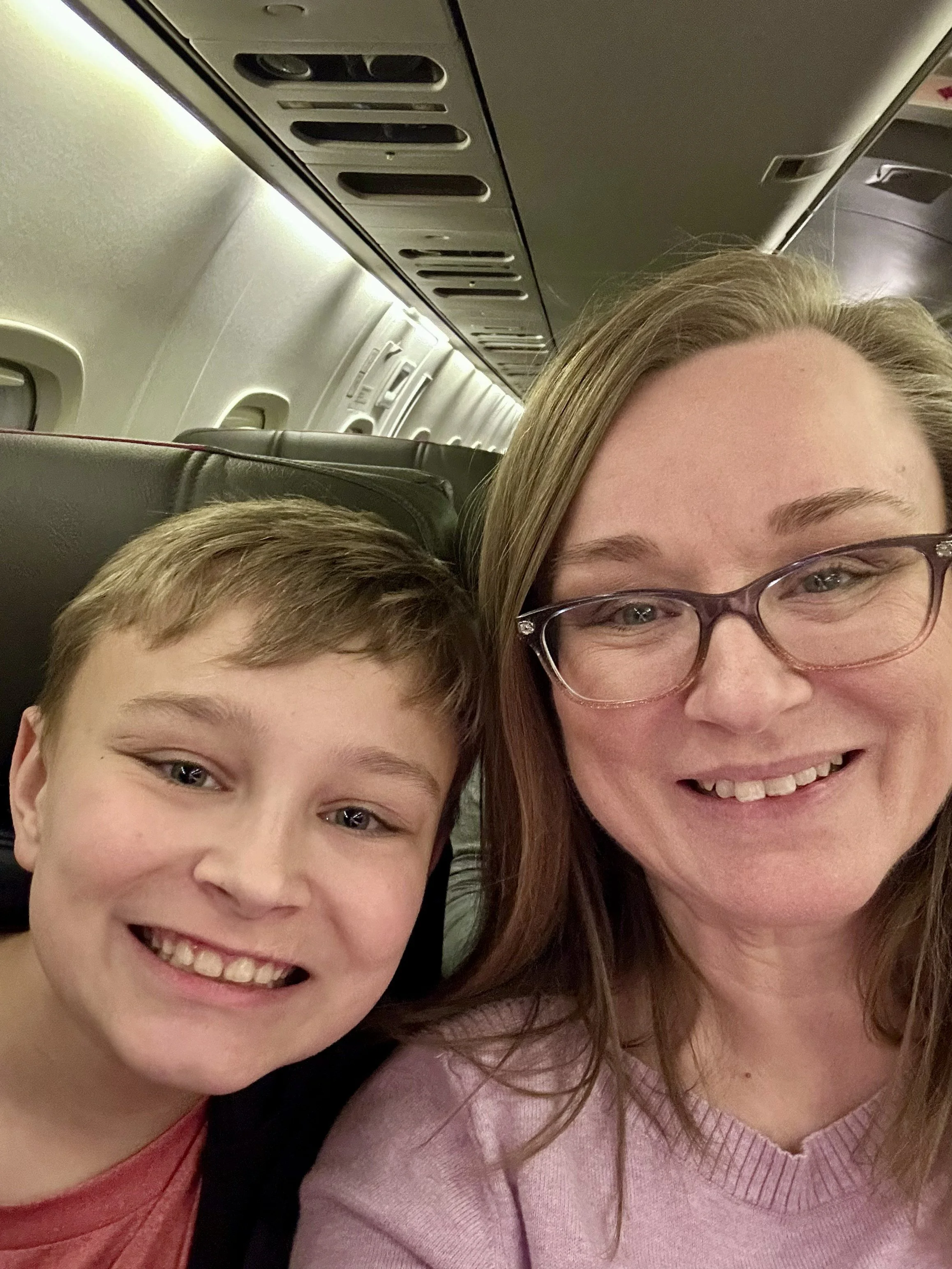 A woman with glasses smiling next to a young boy, both sitting on an airplane.