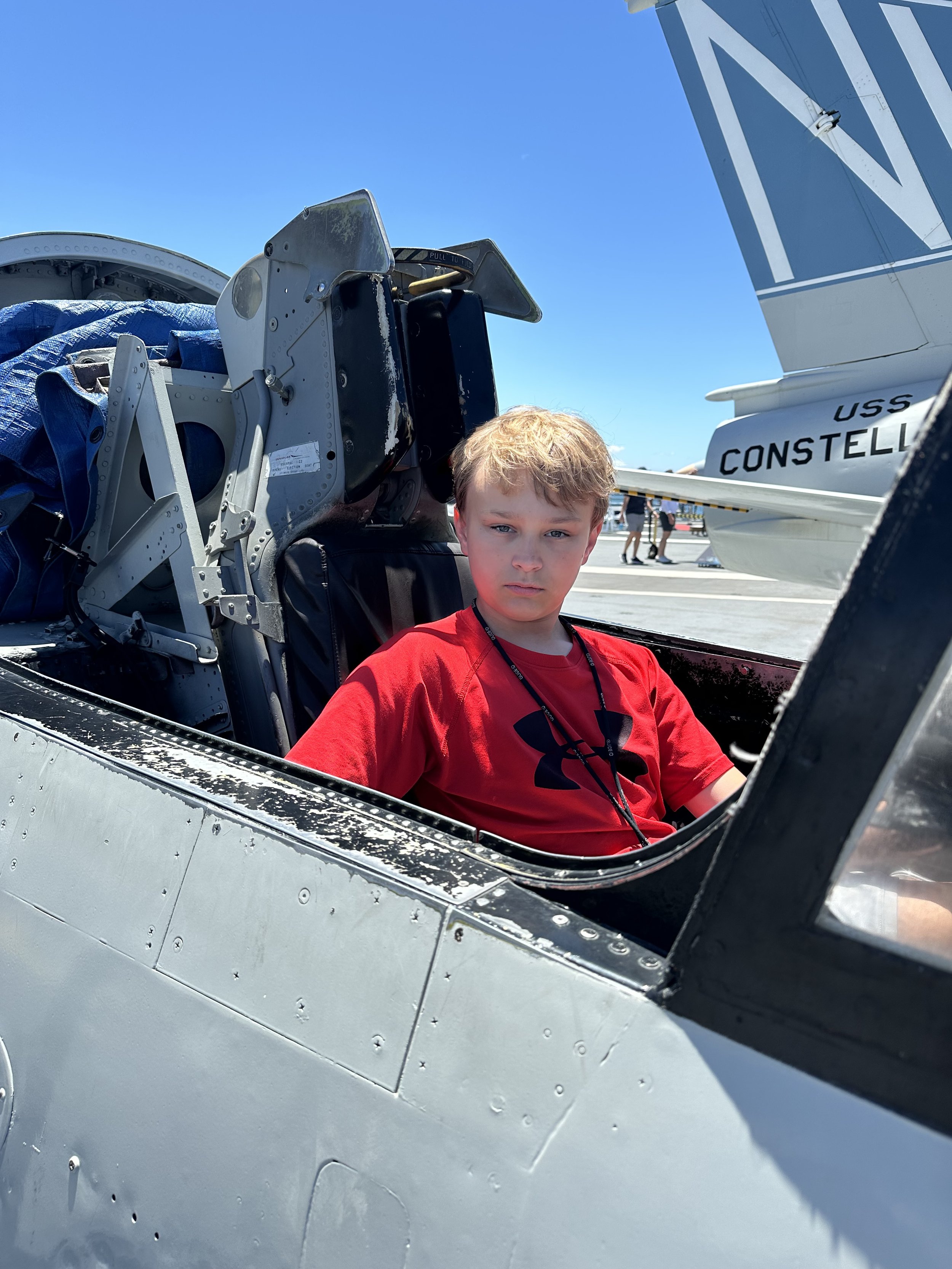 A young boy in a red shirt sitting inside the cockpit of a fighter jet aircraft aboard the Midway in San Diego and part of the jet's tail with the text 'USS CONSTELL' partly visible in the background, under a clear blue sky.
