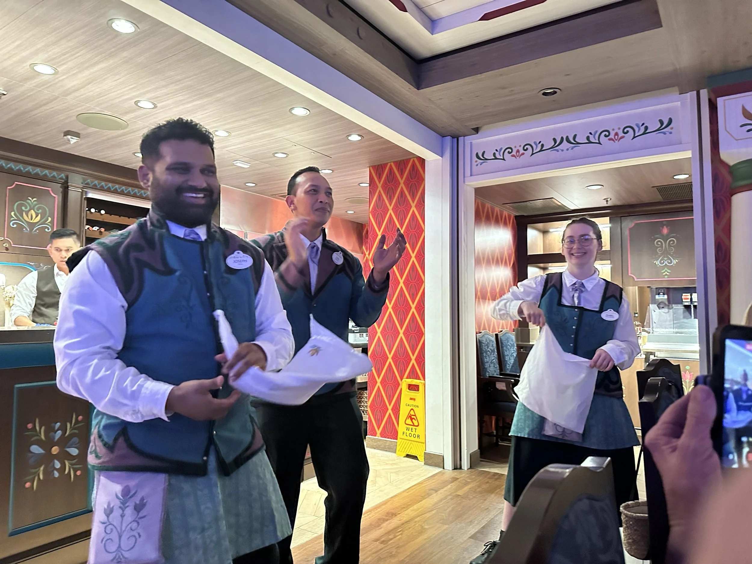 Three restaurant staff members, two men and a woman, standing inside a restaurant, smiling and engaging with each other. The man on the left has a beard and curly hair, wearing a green vest and a white shirt. The man in the middle has short dark hair