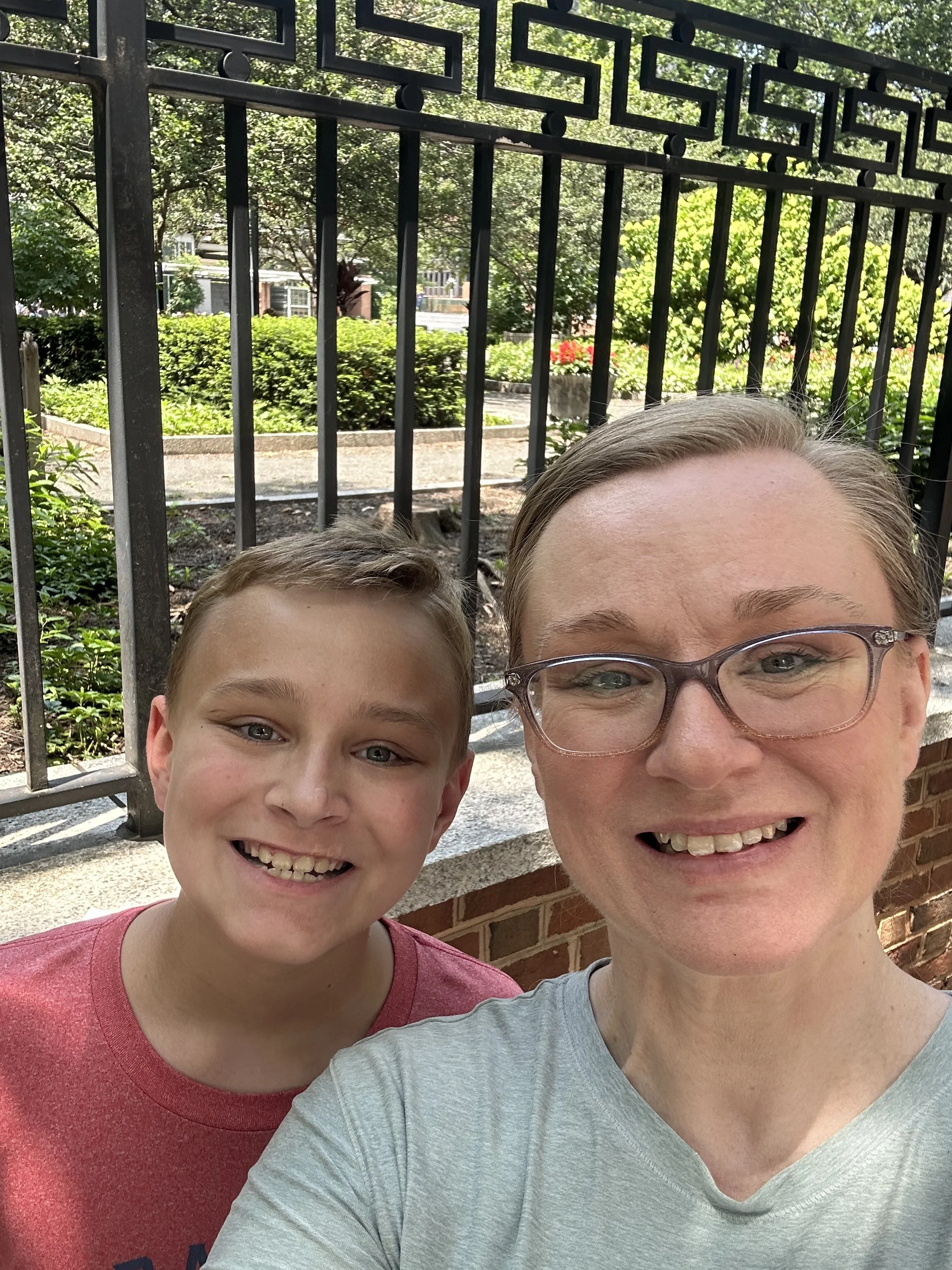 A woman and a boy taking a selfie outdoors in front of a black metal fence with trees and bushes in the background.