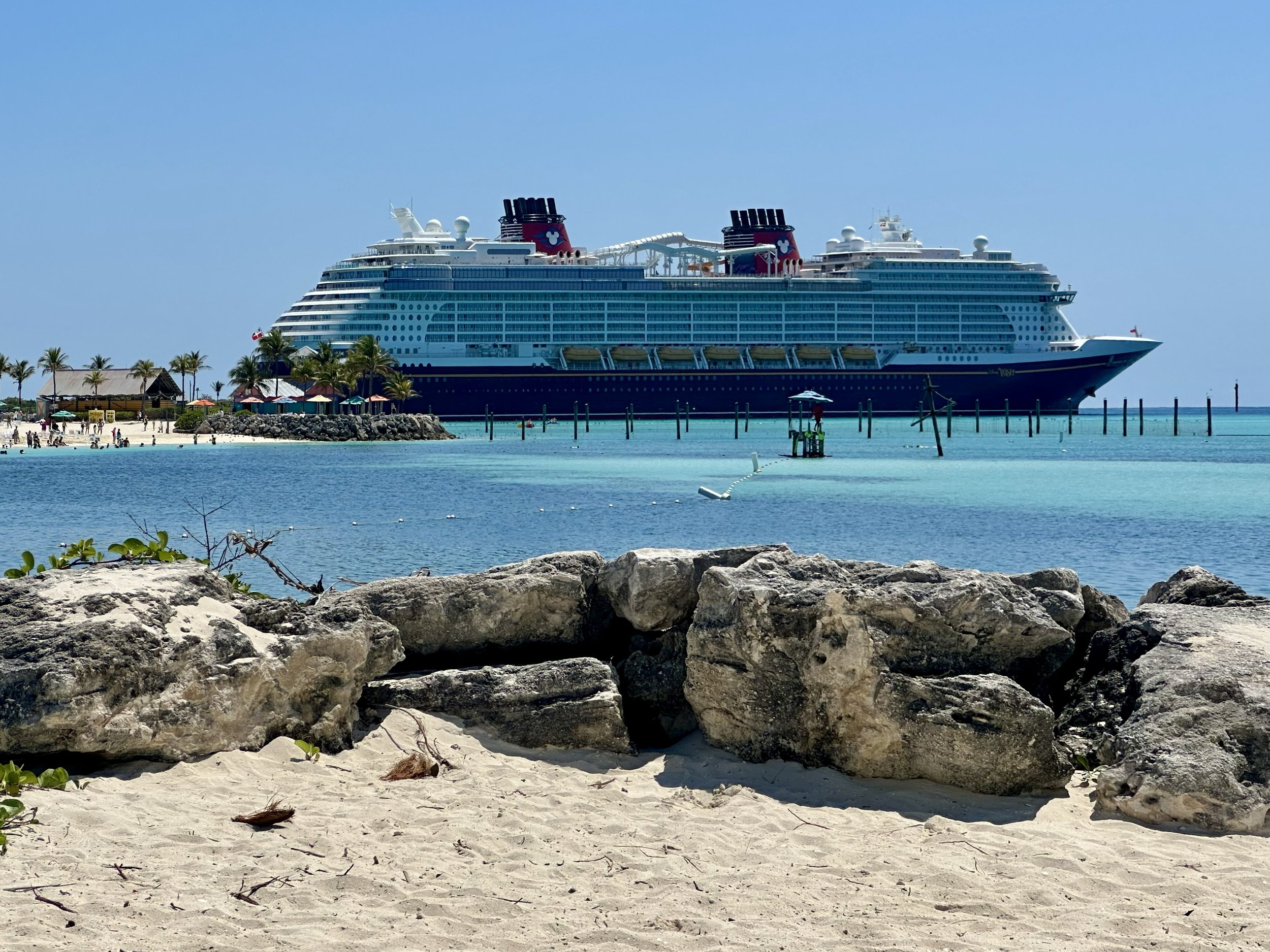 A large cruise ship docked near a sandy beach with rocks, palm trees, and beach umbrellas, against a clear blue sky.