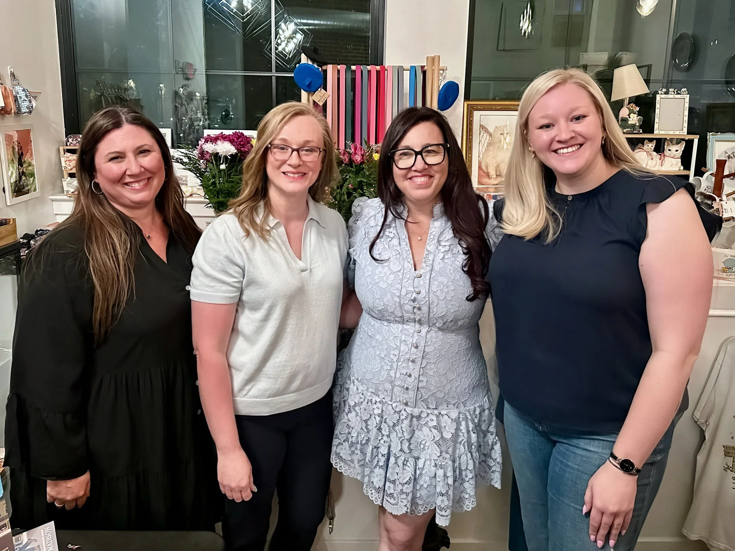 Four women standing close together indoors, smiling. Background includes flowers, artwork, and various decorative items.