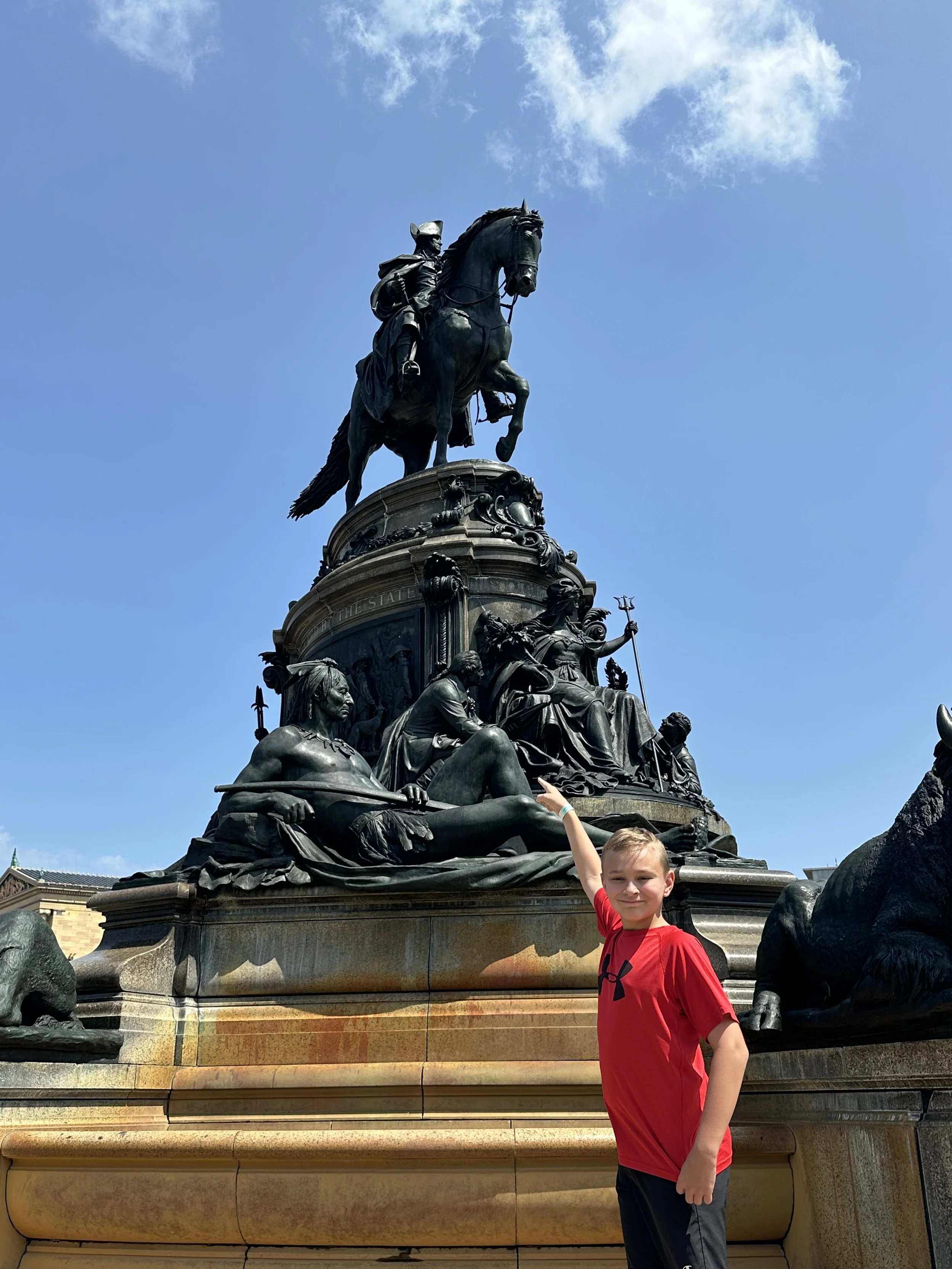 A young boy in a red shirt and black shorts standing in front of Washington monument in Philadelphia with a statue of George Washington on a horse at the top and various other figures at the base, under a partly cloudy blue sky.