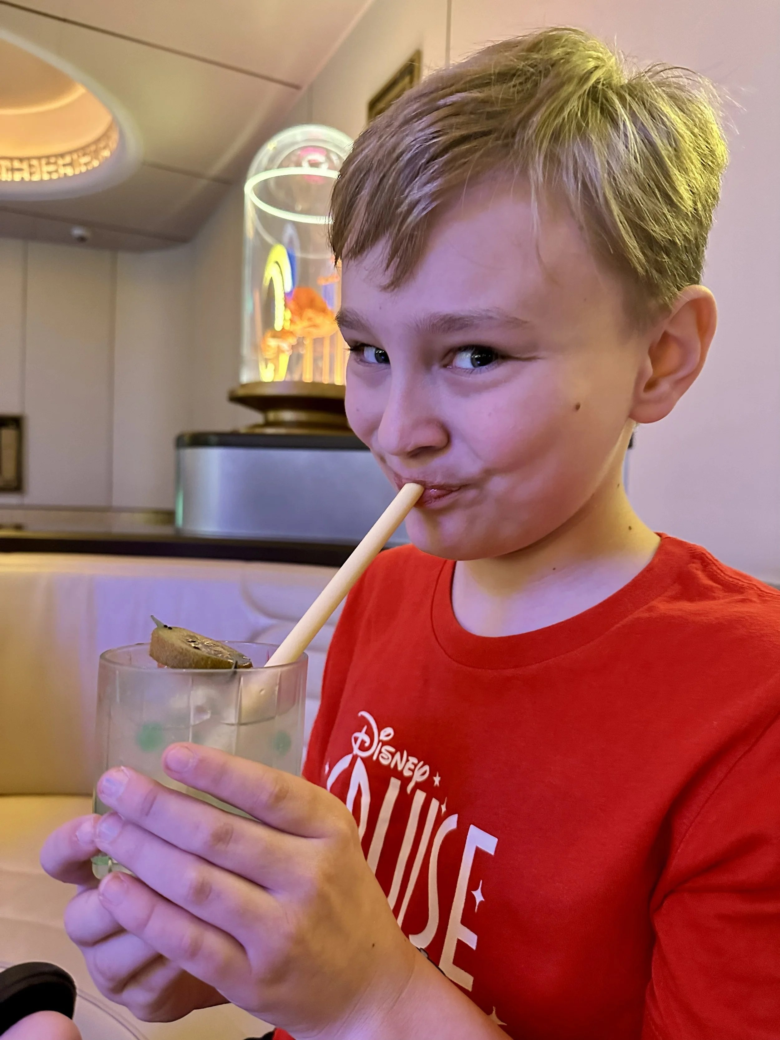 Young boy with blond hair wearing a red Disney T-shirt, holding and drinking from a glass with a straw, in a restaurant or cafe setting.