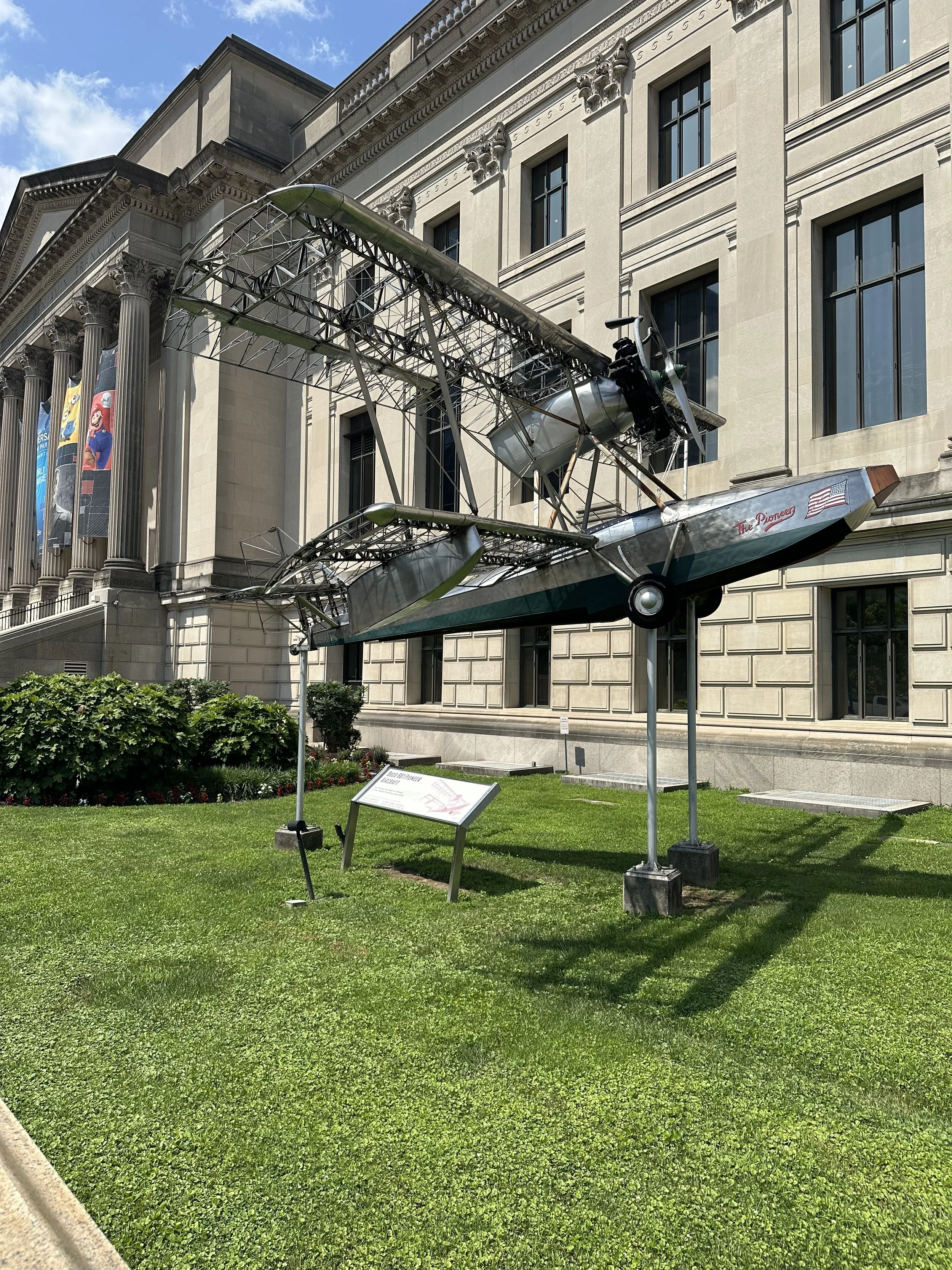 A vintage aircraft sculpture displayed outdoors on a lawn in front of a large, classical-style building with tall windows and decorative columns.
