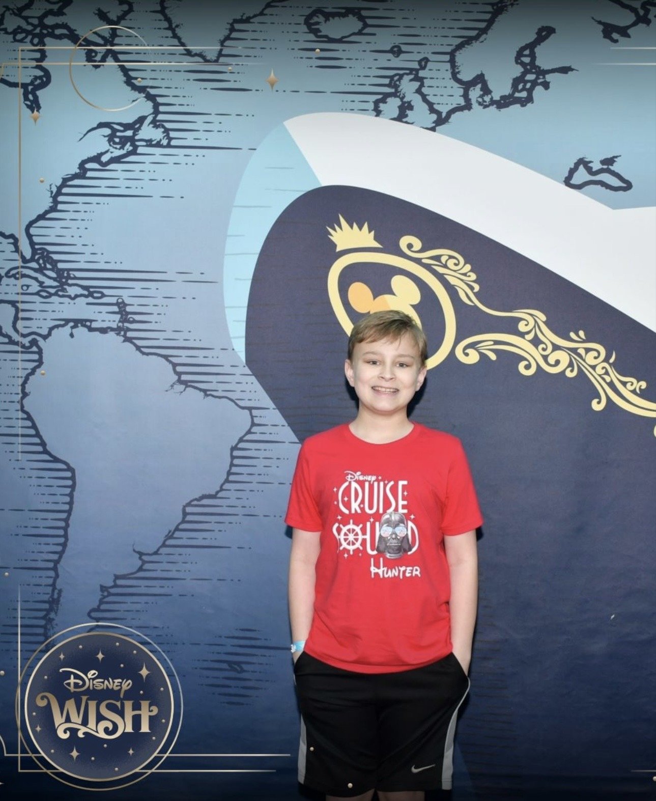 A young boy with short light brown hair smiling, wearing a red Disney Cruise Line T-shirt and black shorts, standing in front of a blue themed backdrop with a world map and Disney Wish logo.