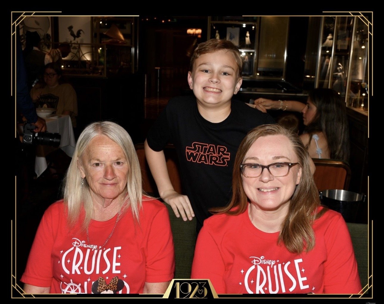 A woman with glasses and a woman with long blonde hair pose for a photo with a boy in a dark Star Wars T-shirt. They are in a dimly lit restaurant or event space. The women wear matching red Disney Cruise shirts, and everyone is smiling.