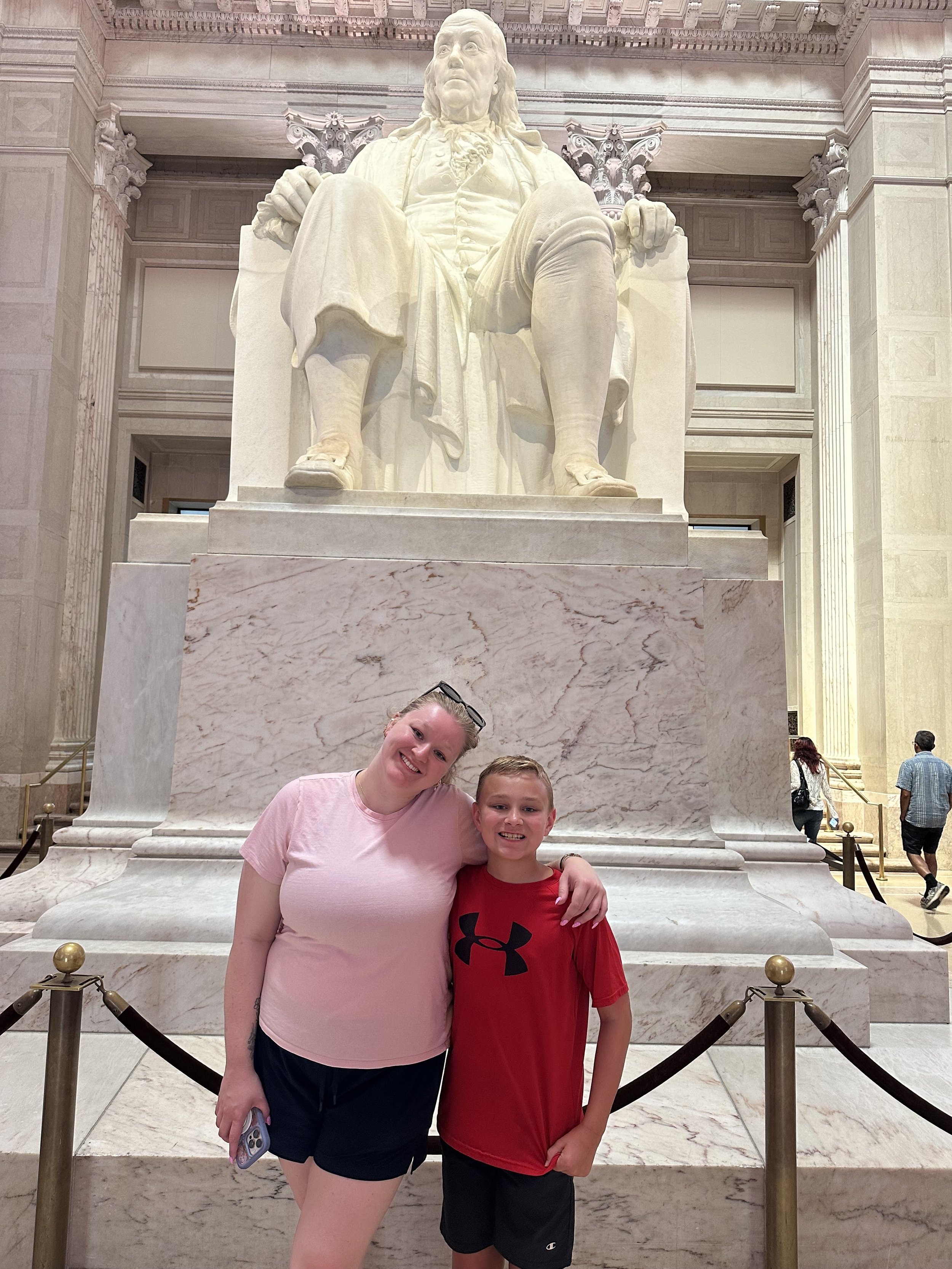A woman and a young boy smiling and posing together in front of the Benjamin Franklin statue at The Franklin Institute in Philadelphia.