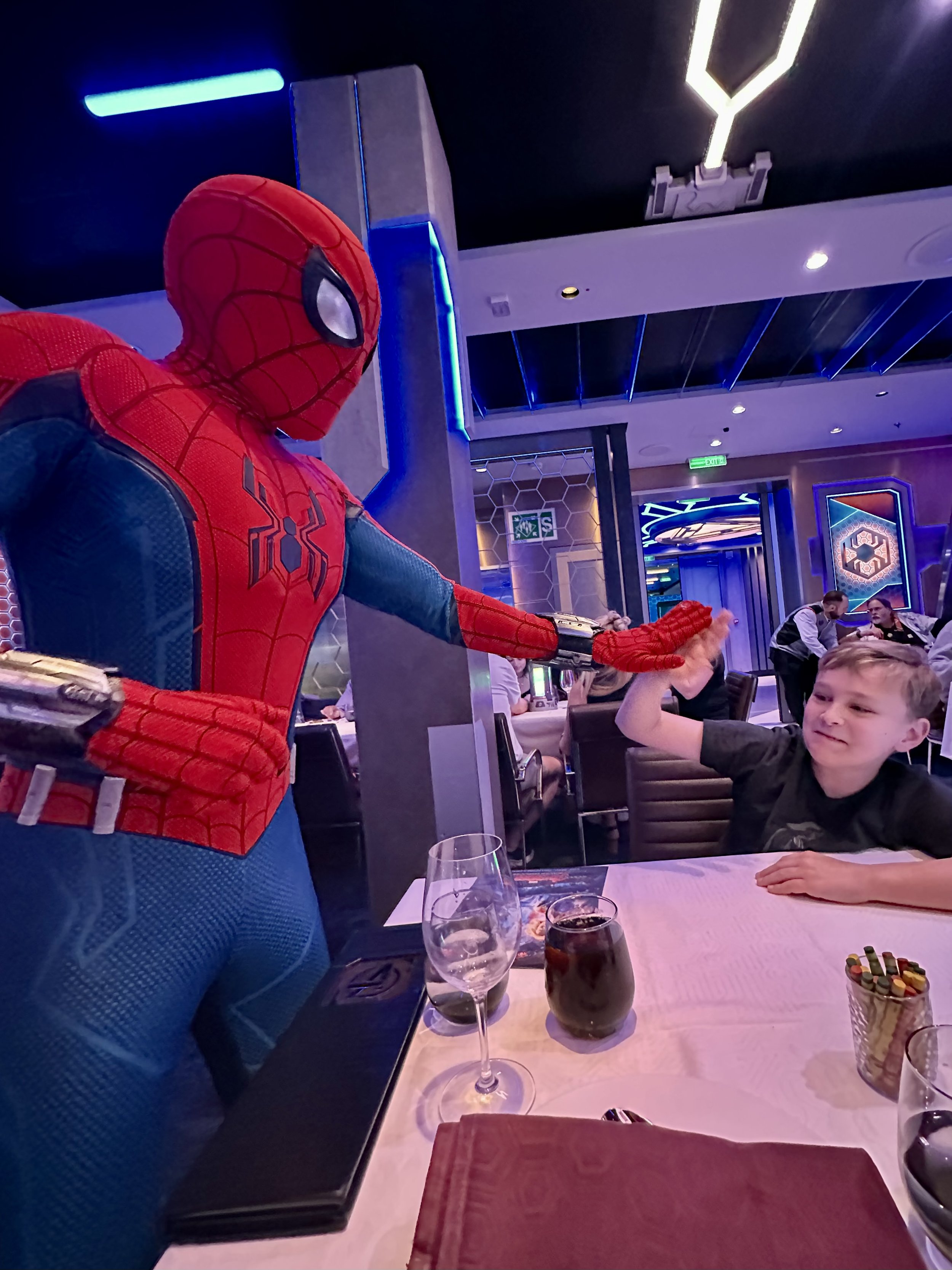 A person dressed as Spider-Man giving a high-five to a young boy at a restaurant table. The table has a glass of red wine, a glass of soda, a box of assorted candies, and a red napkin.