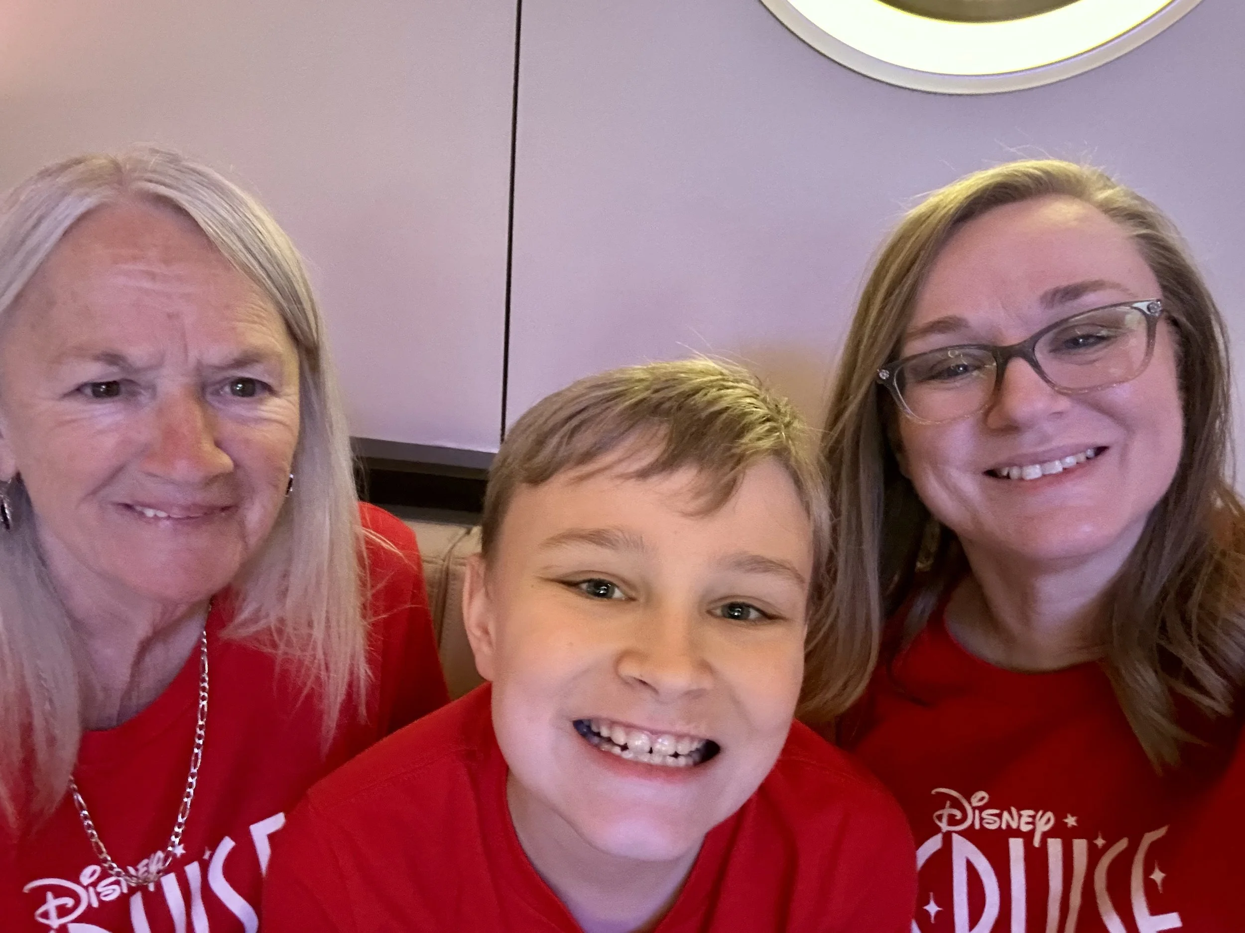 Close-up photo of three smiling people, two women and a young boy, wearing red Disney-themed shirts, in an indoor setting with a purple wall and a round light fixture in the background.