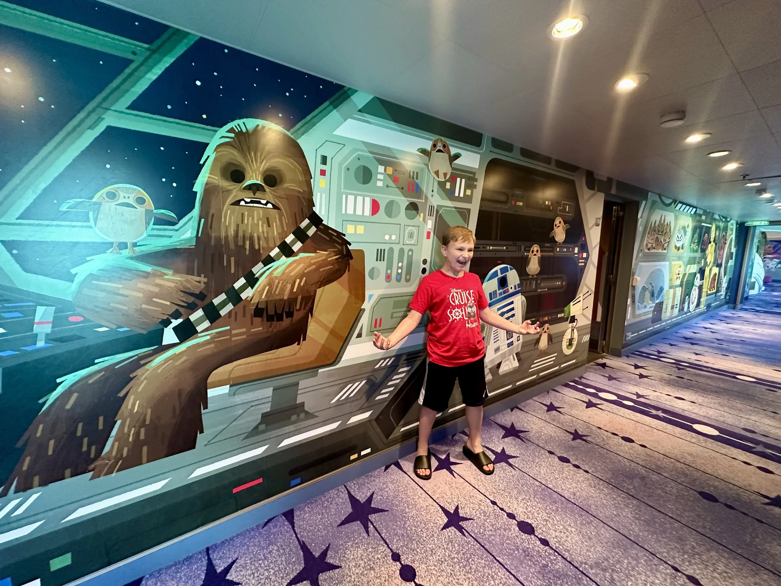 A young boy is standing in front of a colorful, space-themed mural featuring characters from Star Wars, such as Chewbacca and robots, inside a building with lighting on the ceiling and a star-patterned carpet.