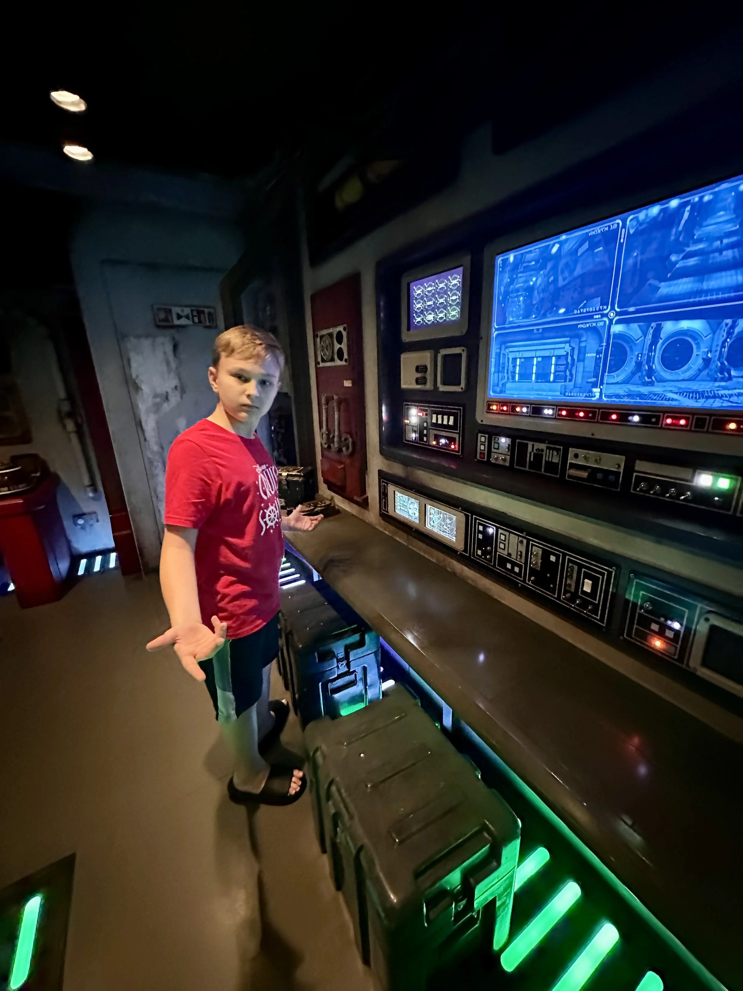 A young boy in a red shirt standing in front of a large computer console with blue digital screens and controls, inside a simulation or control room.