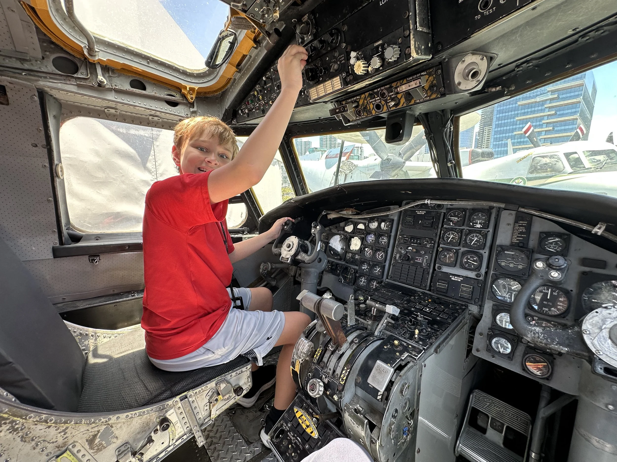 A young boy sitting in the cockpit of a vintage aircraft aboard the Midway, with his right hand on the control stick, surrounded by numerous gauges and switches on the instrument panel, with a city skyline visible through the windows outside.