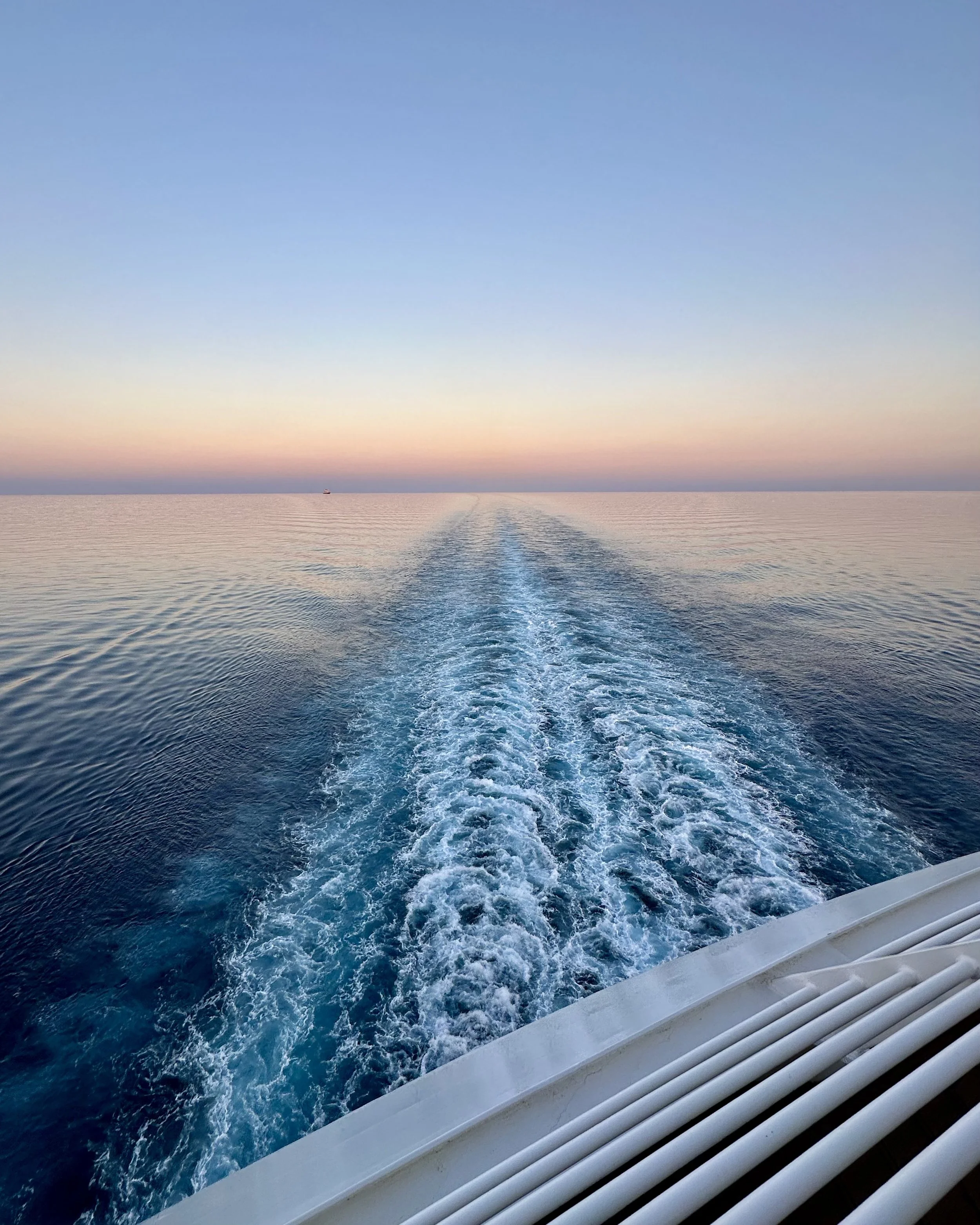 View of the ocean with a boat in the distance and a boat wake trail in the water during sunset or sunrise, with a window frame with air vents in the foreground.