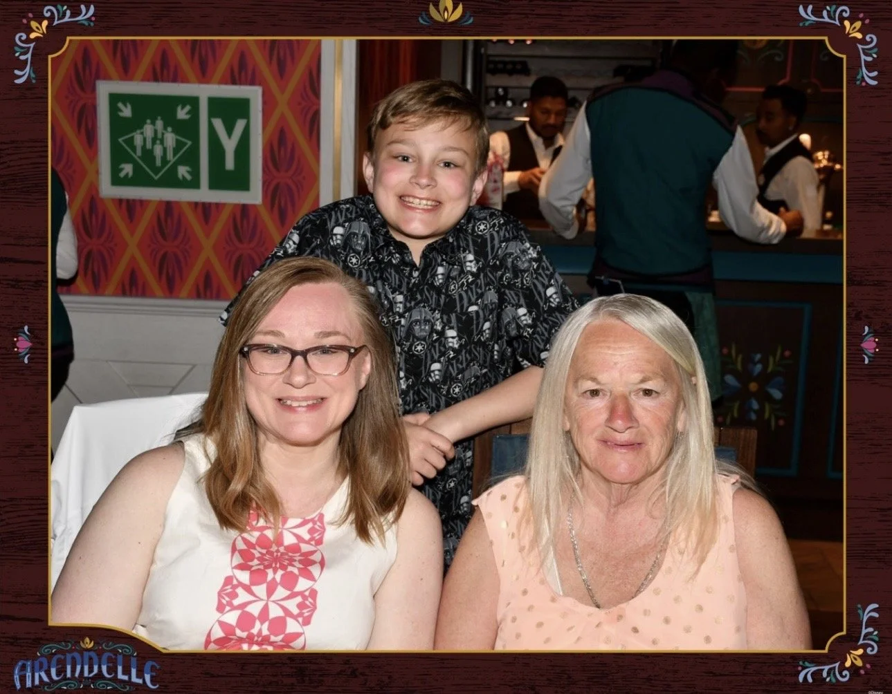 A smiling boy standing behind two women, one with glasses, in a restaurant with waitstaff in the background.