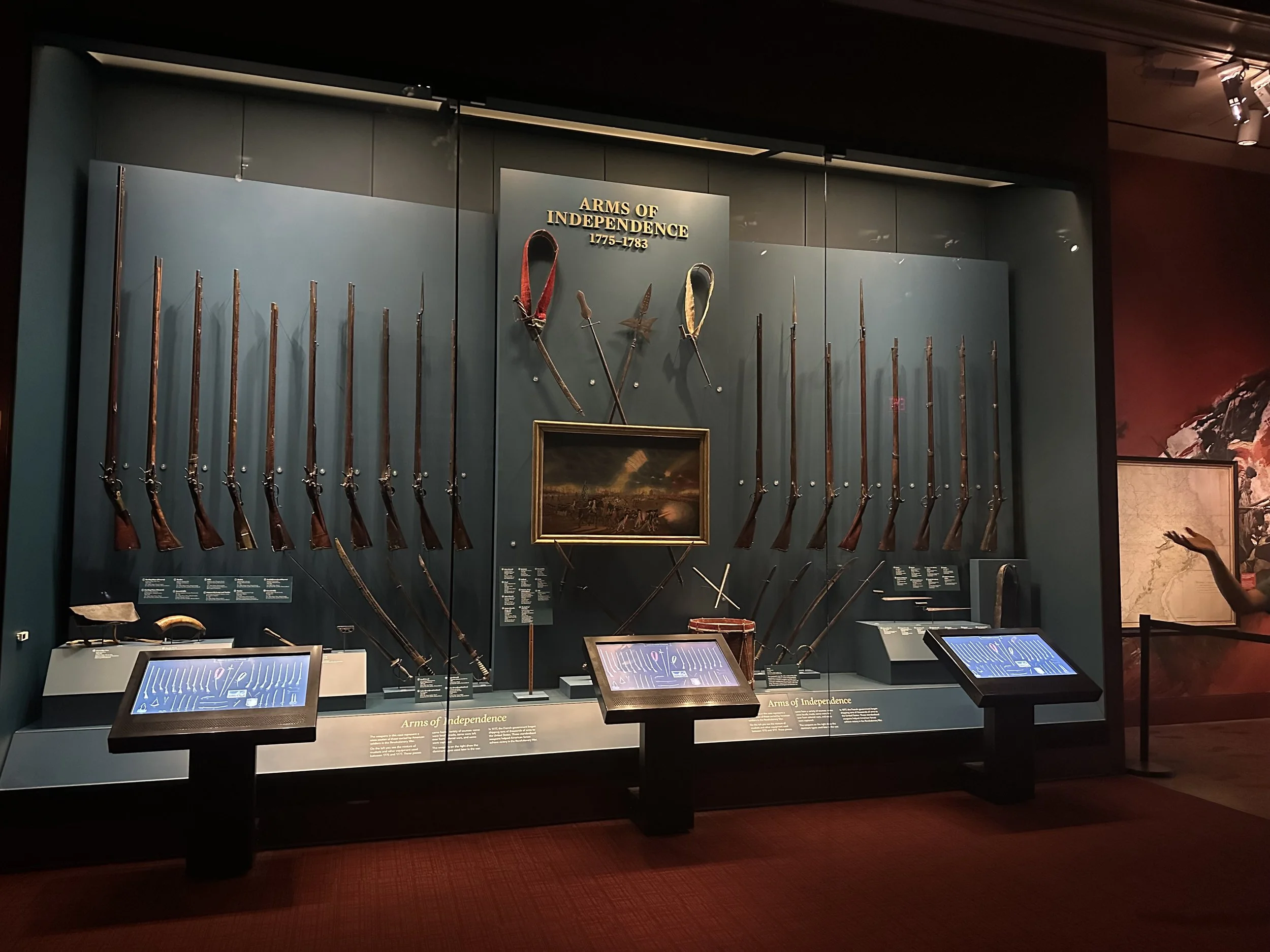 Display case titled "Arms of Independence 1775-1783" showing various historical weapons, including muskets and swords, with informative placards and a painting underneath.