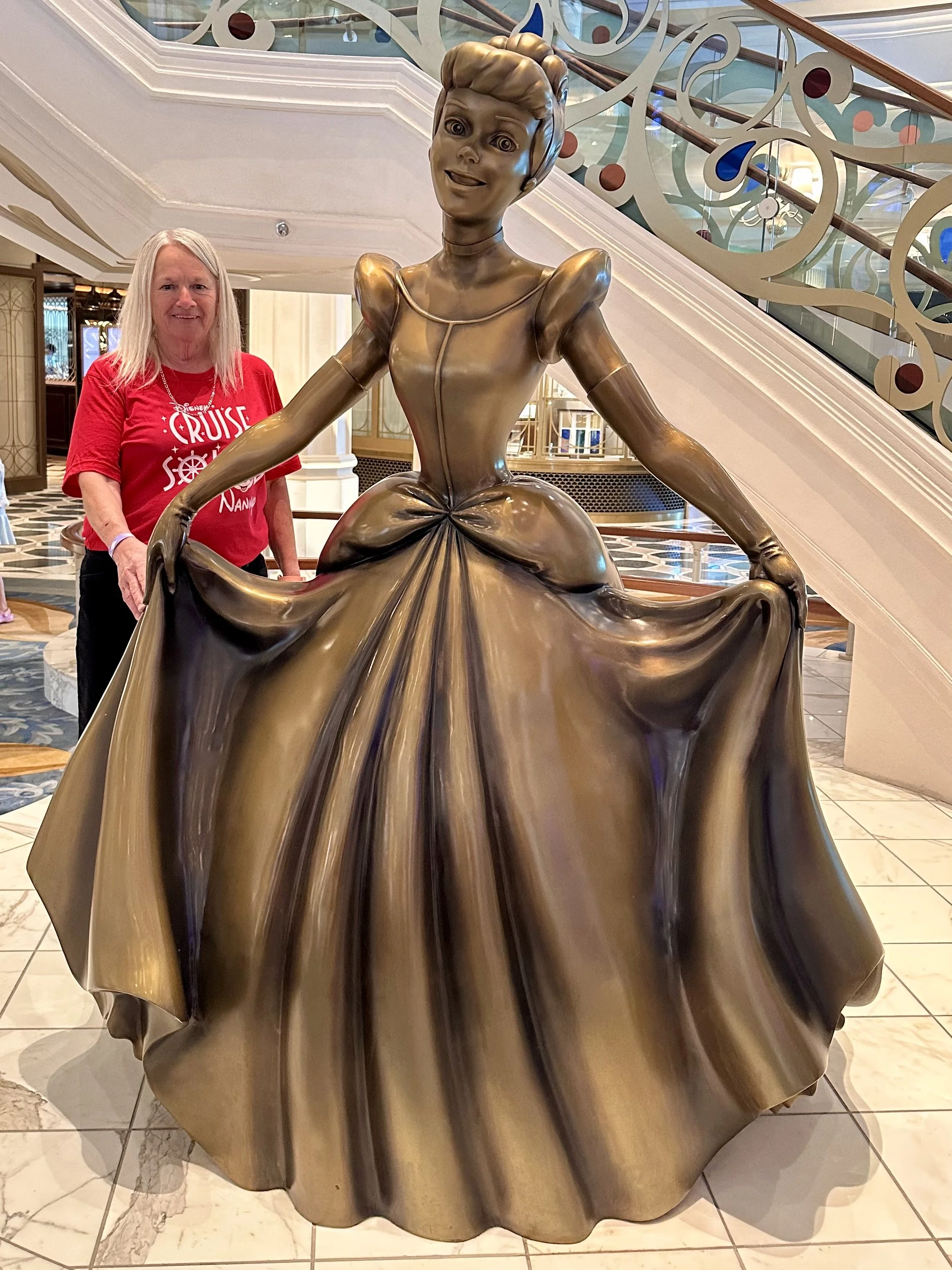 A woman standing next to a golden statue of a princess in a large, ornate hotel lobby. The statue depicts a young woman in a flowing gown, holding the skirt with both hands, with an elaborate hairstyle. The woman is wearing a red shirt with white tex