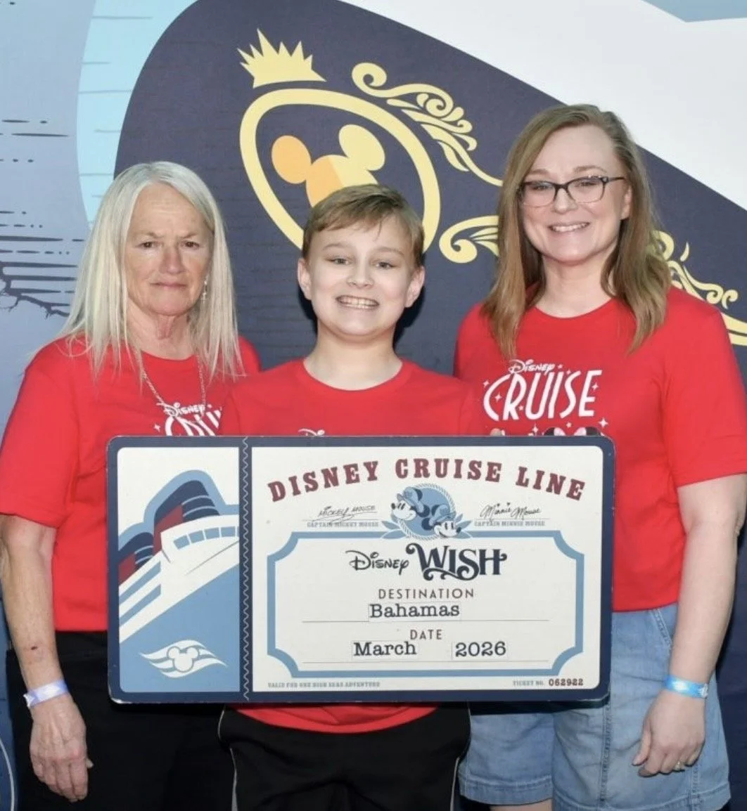 Three people holding a large Disney Cruise Line ticket, with a woman and girl on the right smiling and a woman on the left with a serious expression, all wearing red Disney Cruise Line shirts, standing in front of a themed backdrop.