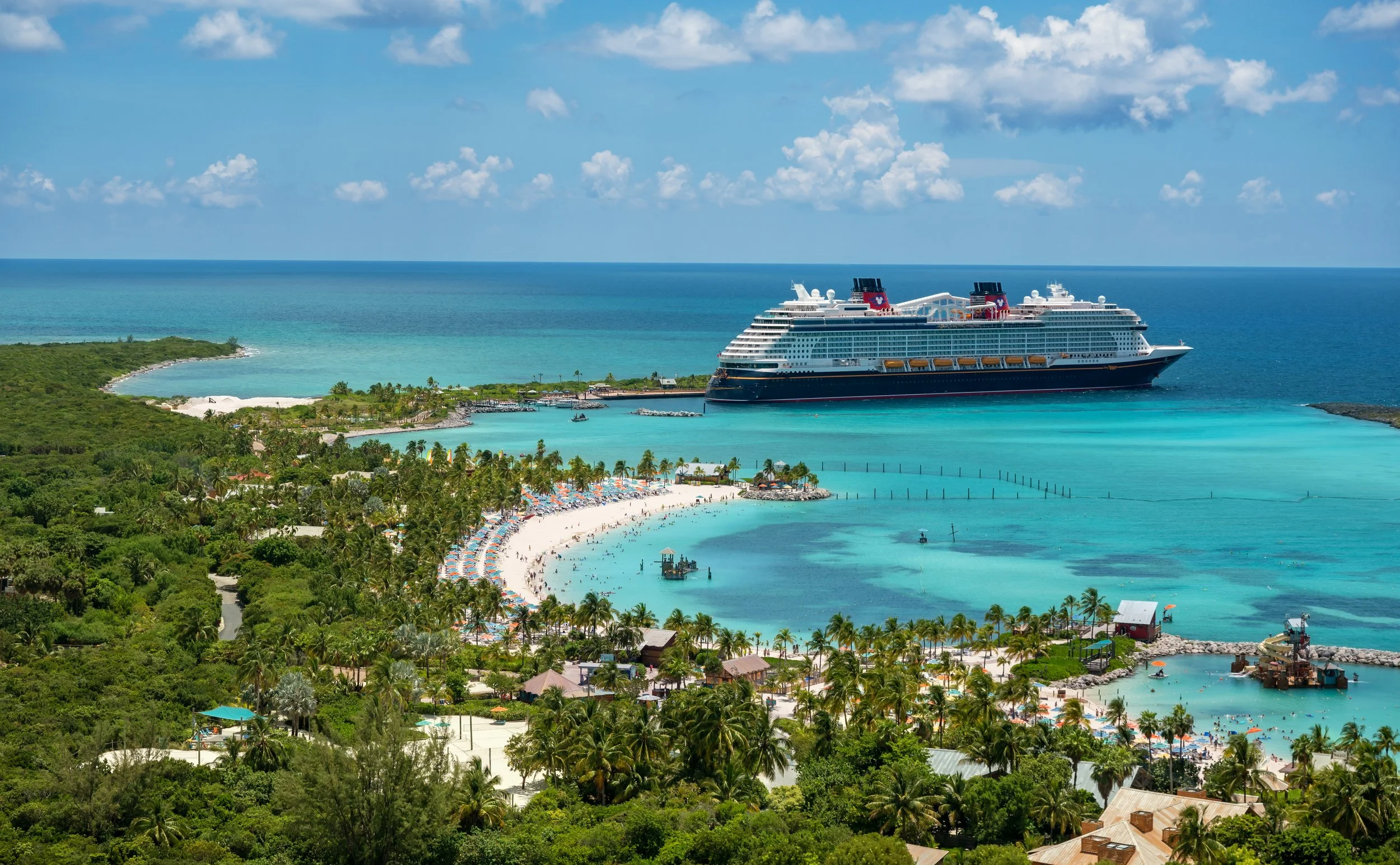 A large cruise ship docked near a tropical island with clear blue waters, a sandy beach with umbrellas, and green lush vegetation, under a partly cloudy sky.