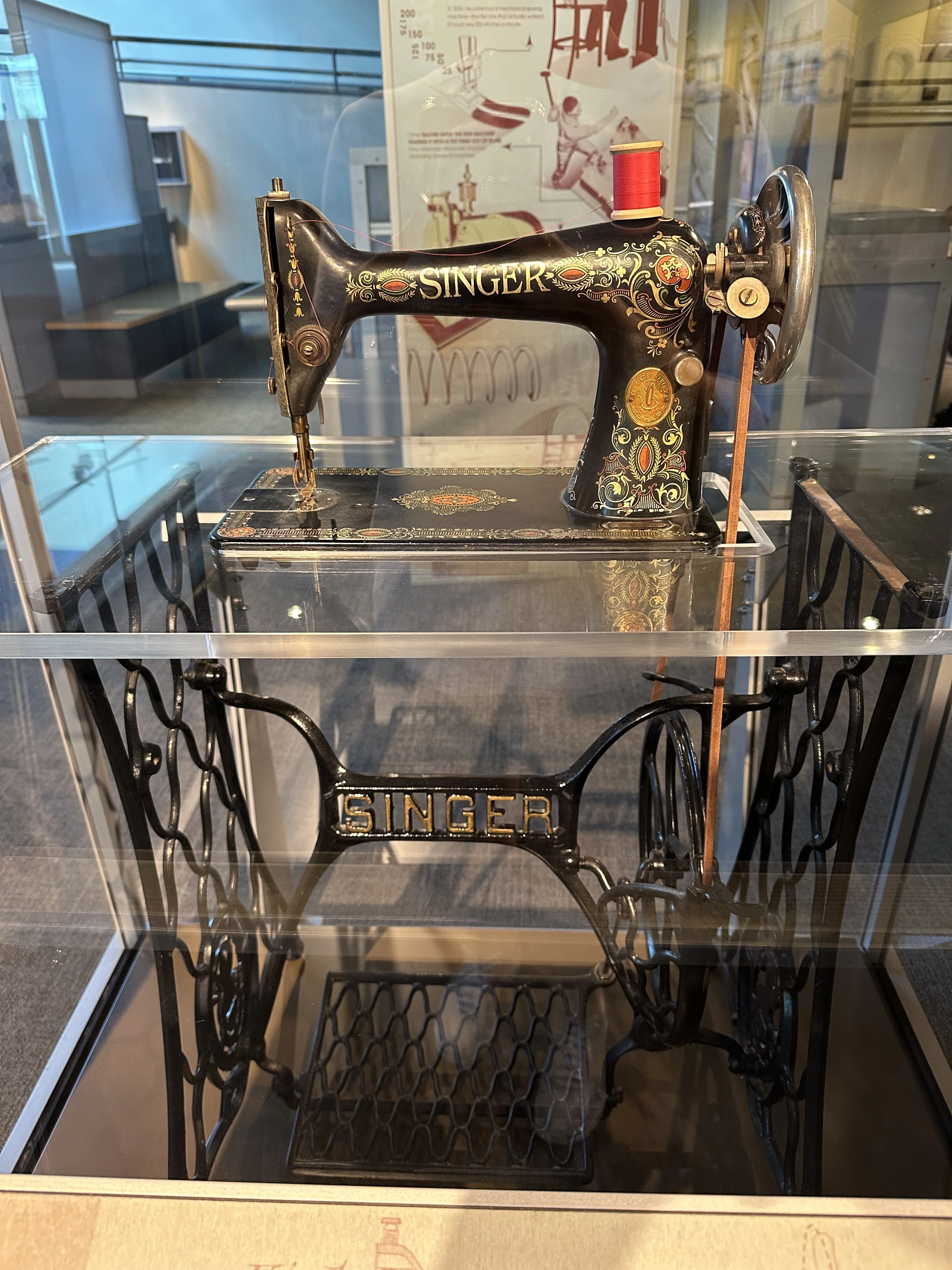 Vintage black sewing machine with decorative gold and orange patterns, labeled 'SINGER', displayed in a glass case.