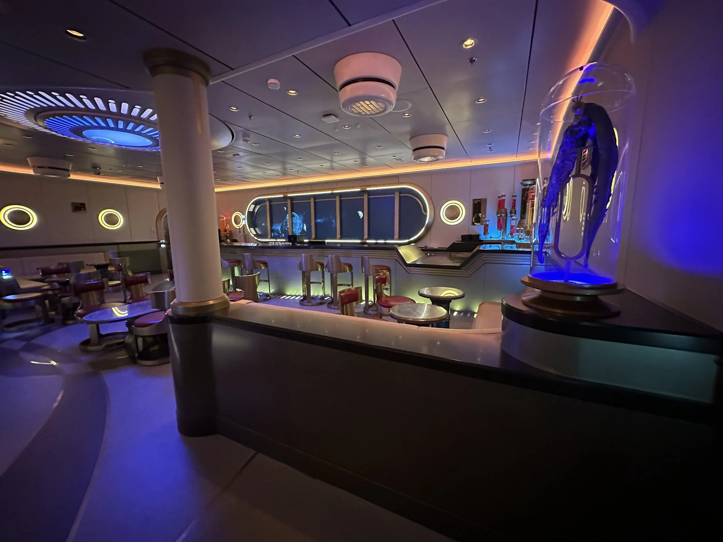 Interior of a space-themed lounge with futuristic decor, round wall lights, a window showing space and planets, and a bright blue-lit display in a transparent case.