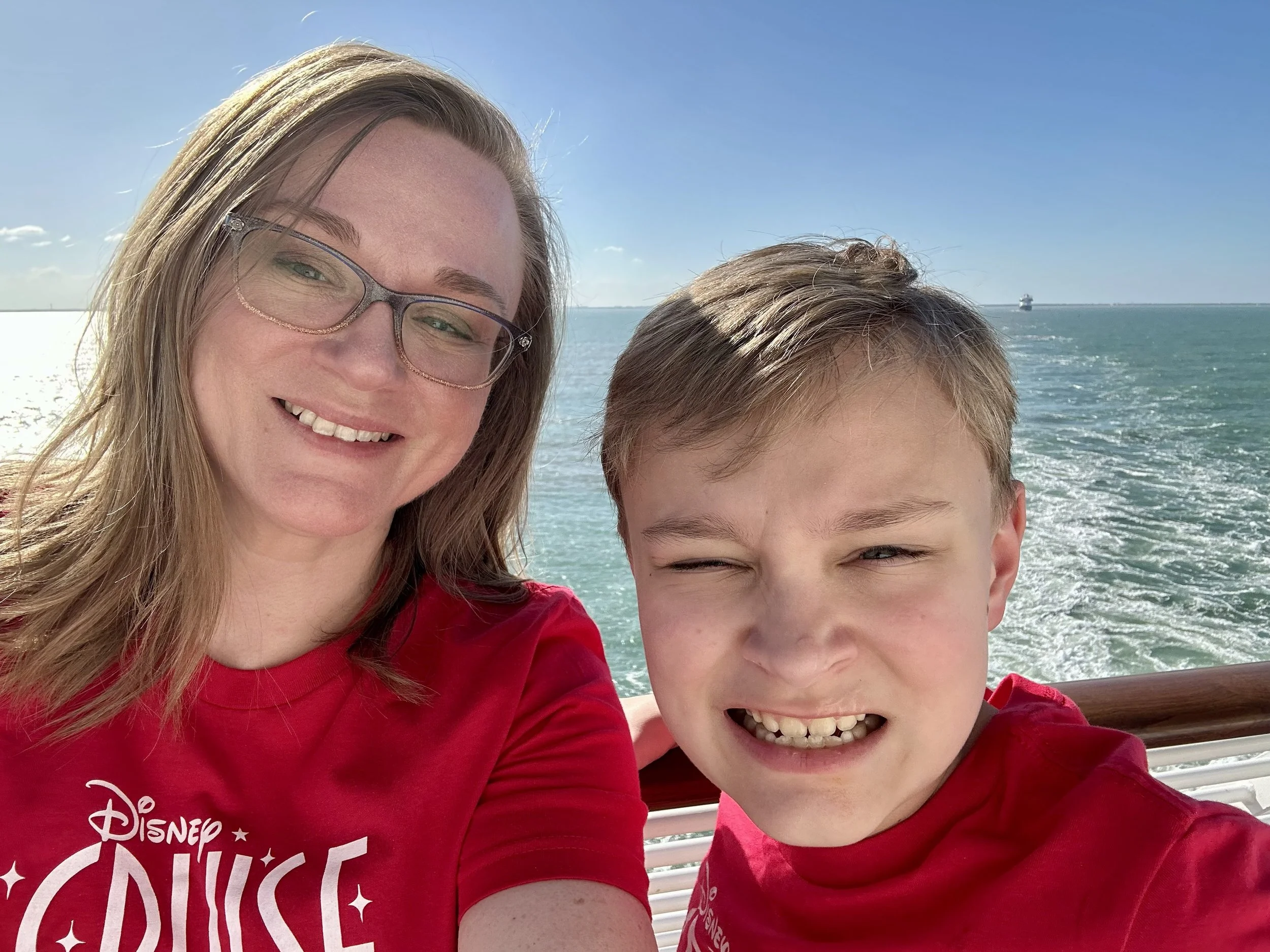 A woman and a young boy smiling for a selfie on a boat with the ocean and a ship in the background.