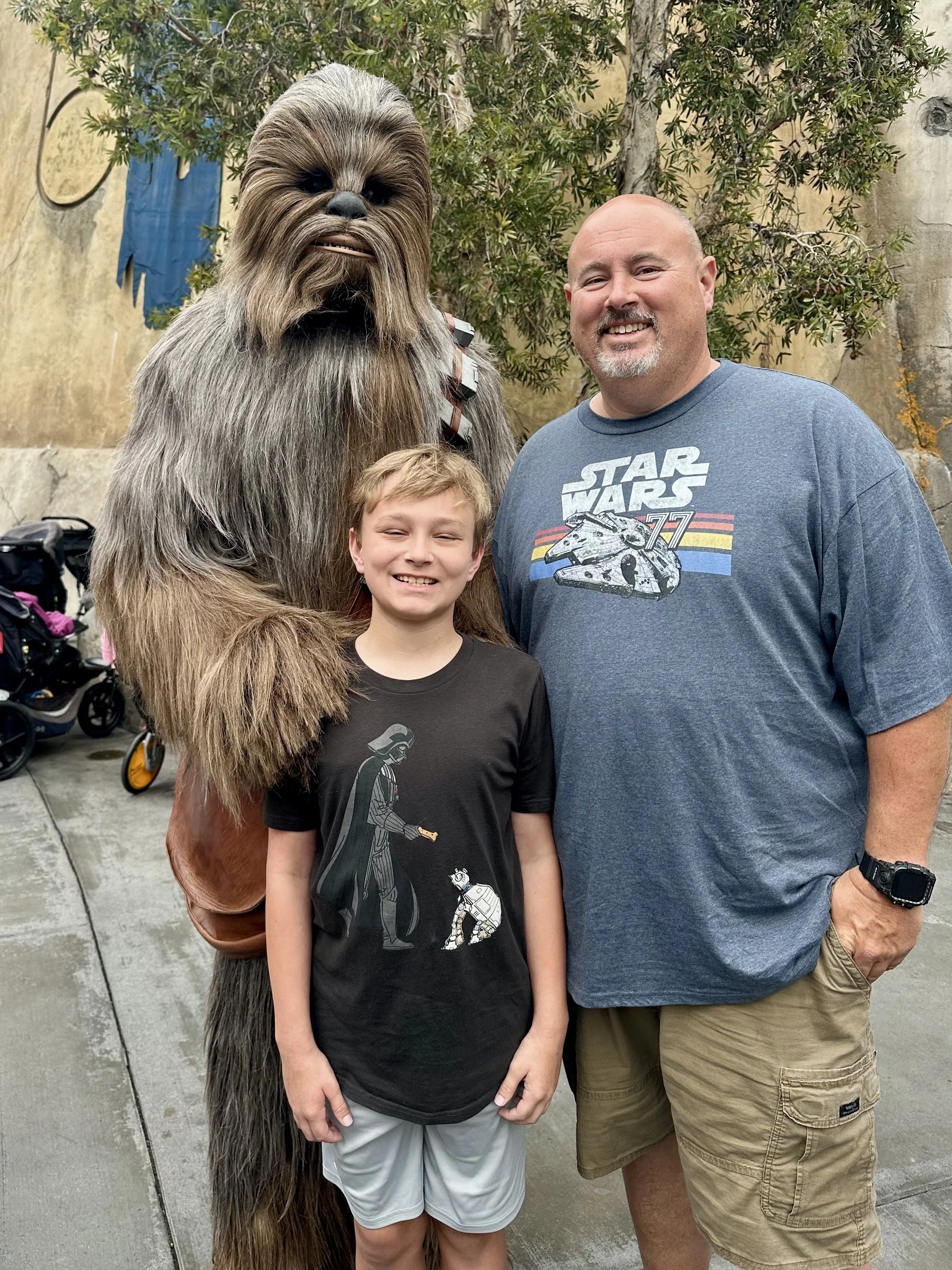 A person dressed as Chewbacca from Star Wars standing between a young boy and an adult man, both smiling, outdoors with trees and a beige wall in the background.