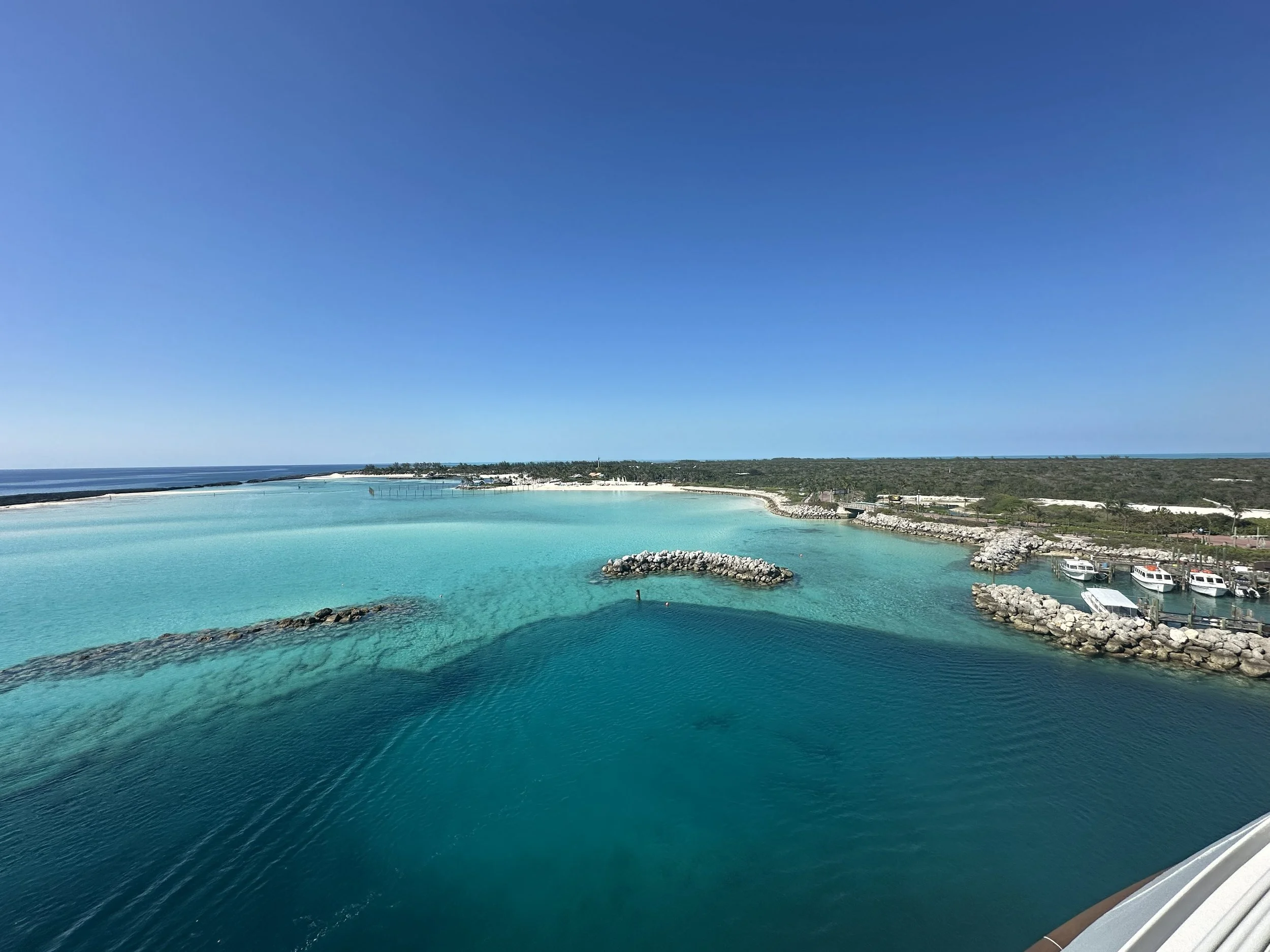 A tropical coastal scene with turquoise waters, a sandy beach, and a breakwater. There are boats docked in the harbor and a distant landmass with trees under a clear blue sky.