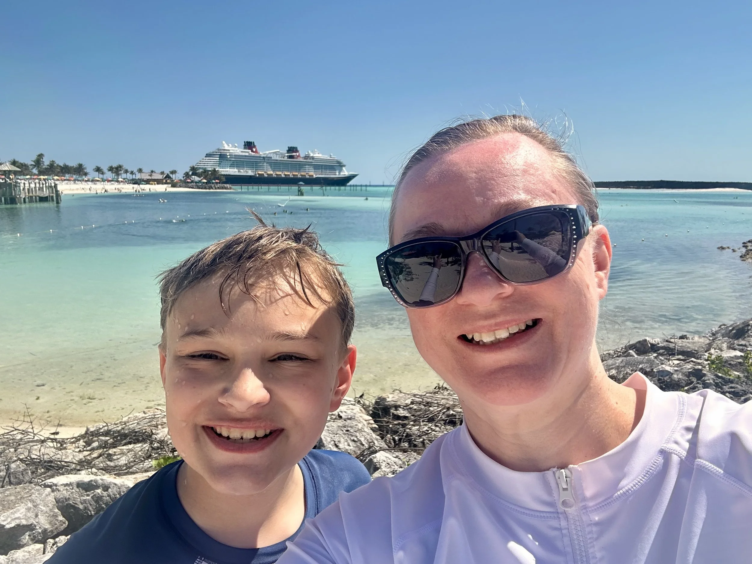 A woman and a young boy are taking a selfie at the beach. Behind them, the ocean, a cruise ship, and palm trees are visible under a clear blue sky.