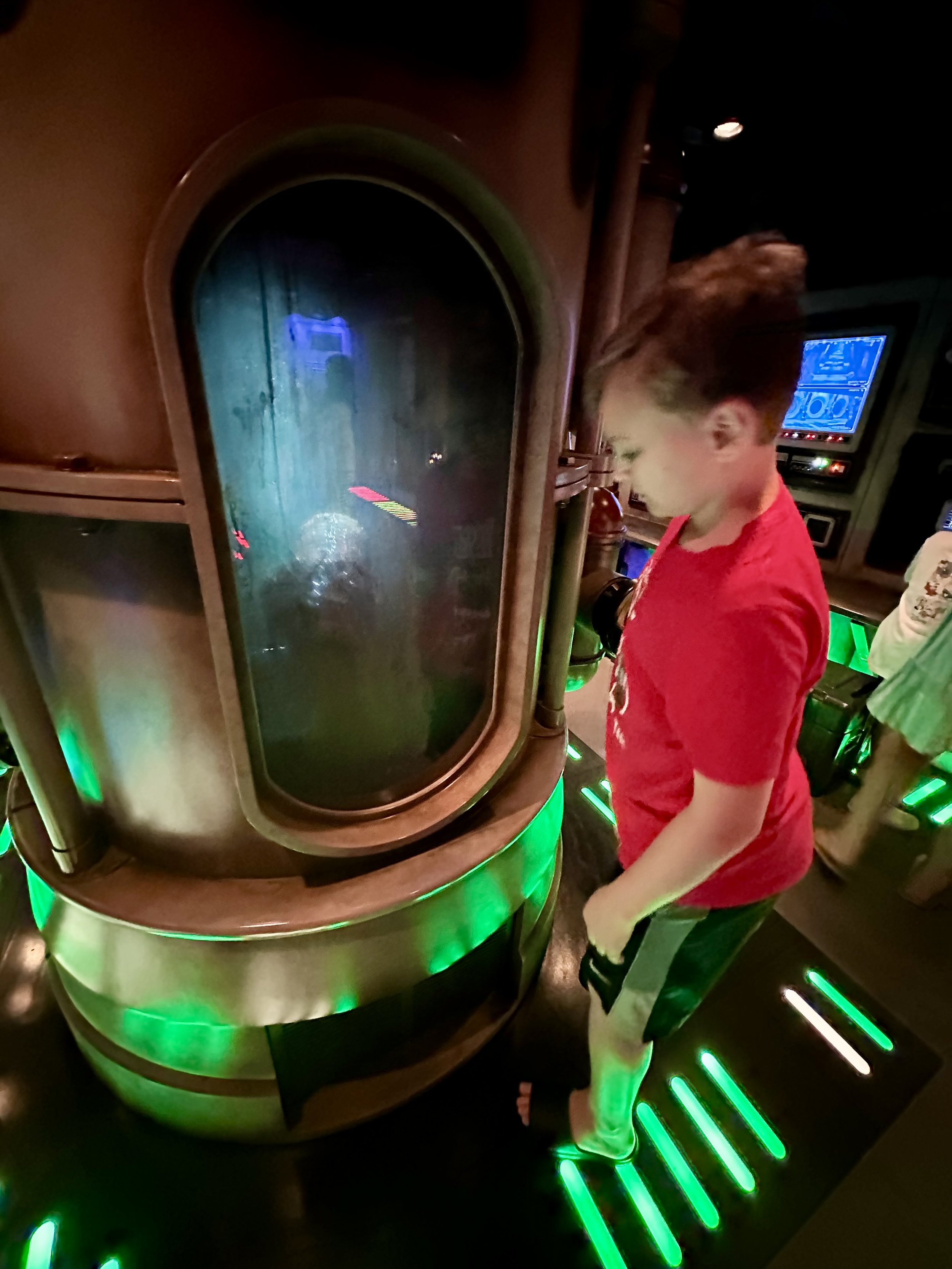 A young boy in a red shirt and shorts stands in front of a sci-fi style, green-lit control station with a large, glass display case filled with water and objects inside, in an indoor arcade or entertainment venue.