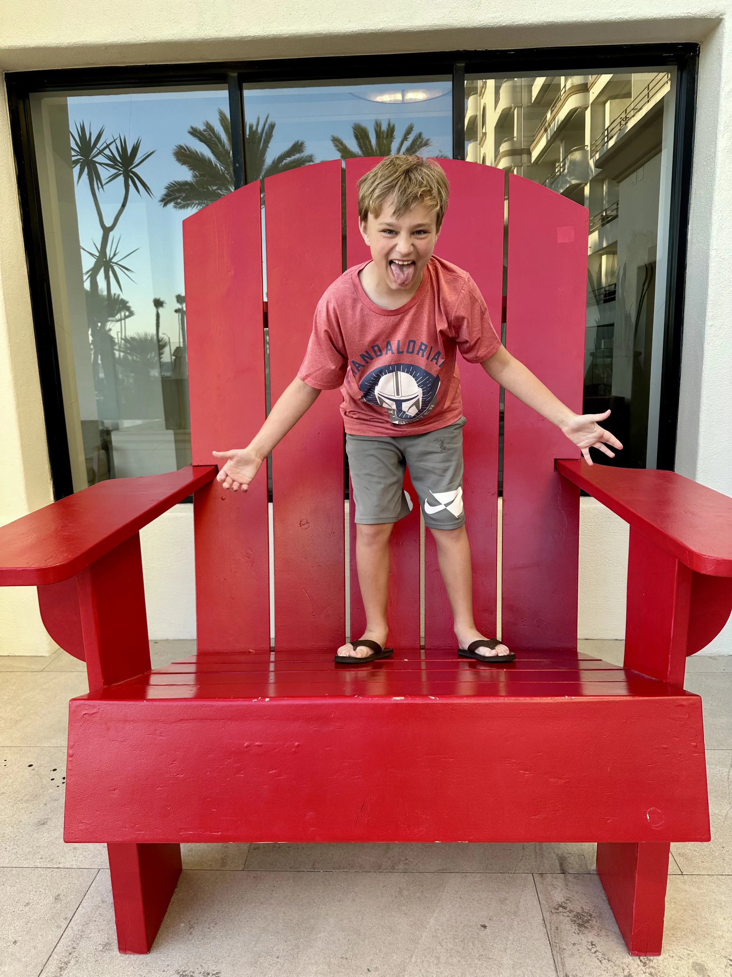 A young boy standing on a large red chair with a backrest that resembles a giant Adirondack chair, sticking out his tongue and making a playful gesture with his arms.
