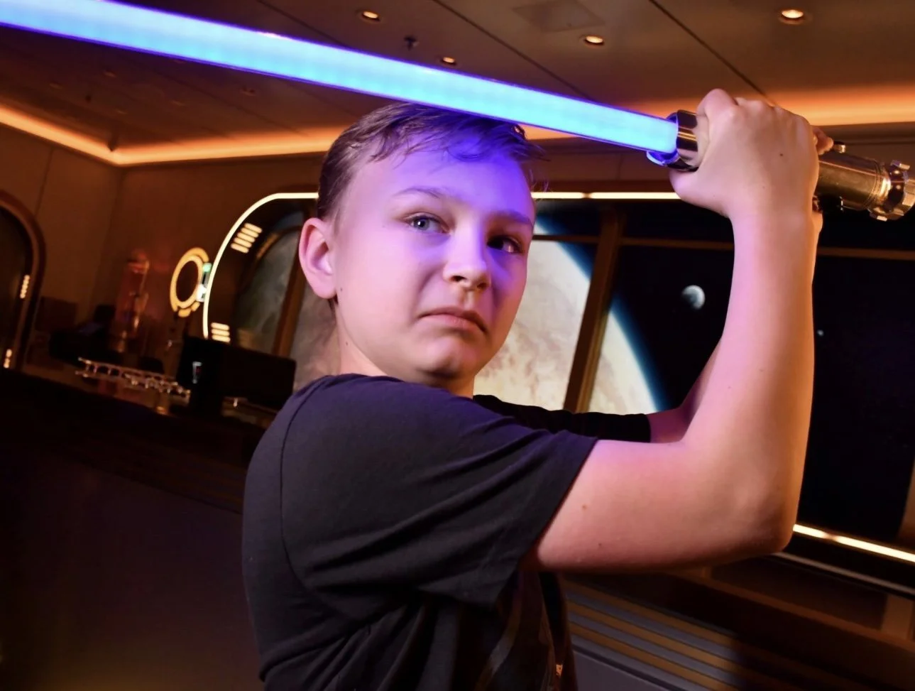 Young person holding a blue lightsaber toy in space-themed room with planet and moon images on the background.