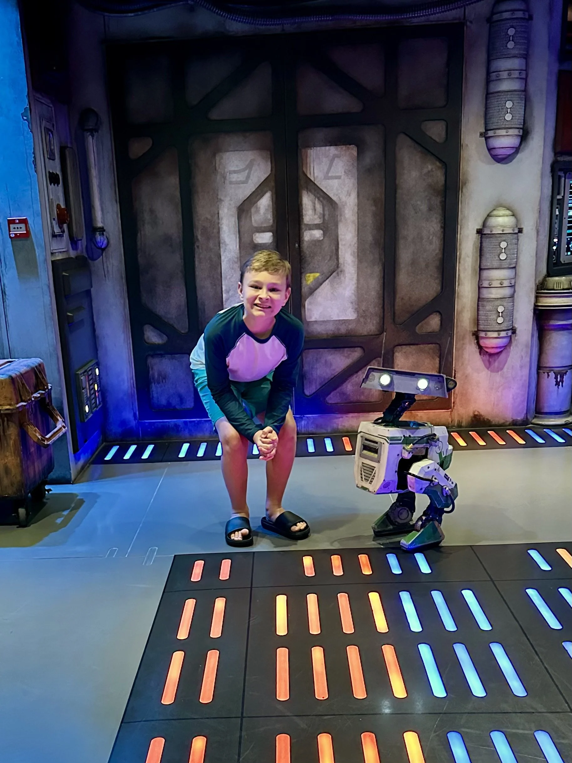 A young boy with a scrunched face kneeling in front of a robot, inside a sci-fi themed room with illuminated lines on the floor and futuristic wall designs.