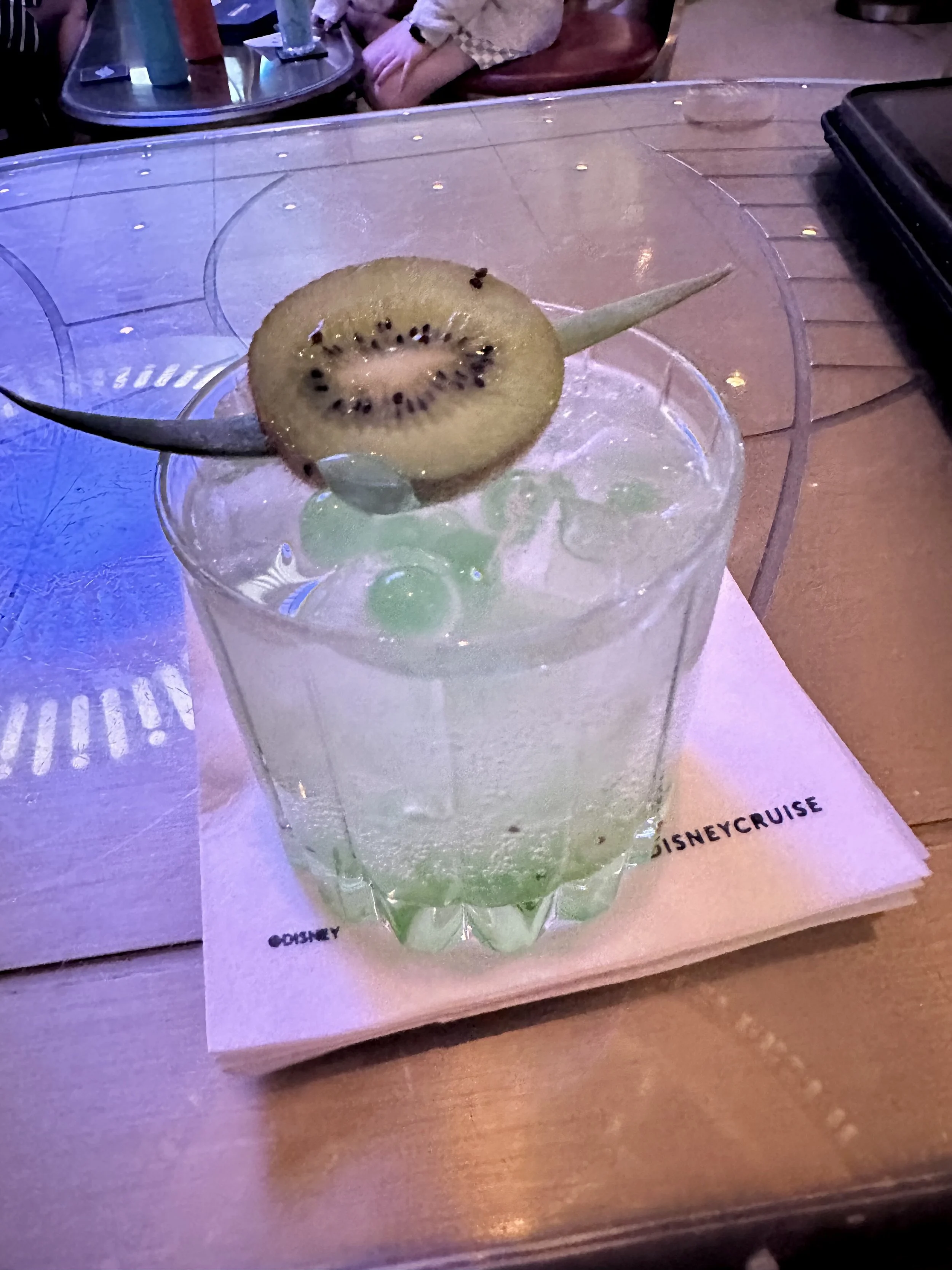 A glass of green-colored drink with a slice of kiwi on the rim. The drink sits on a Disney-themed place mat on a wooden table.