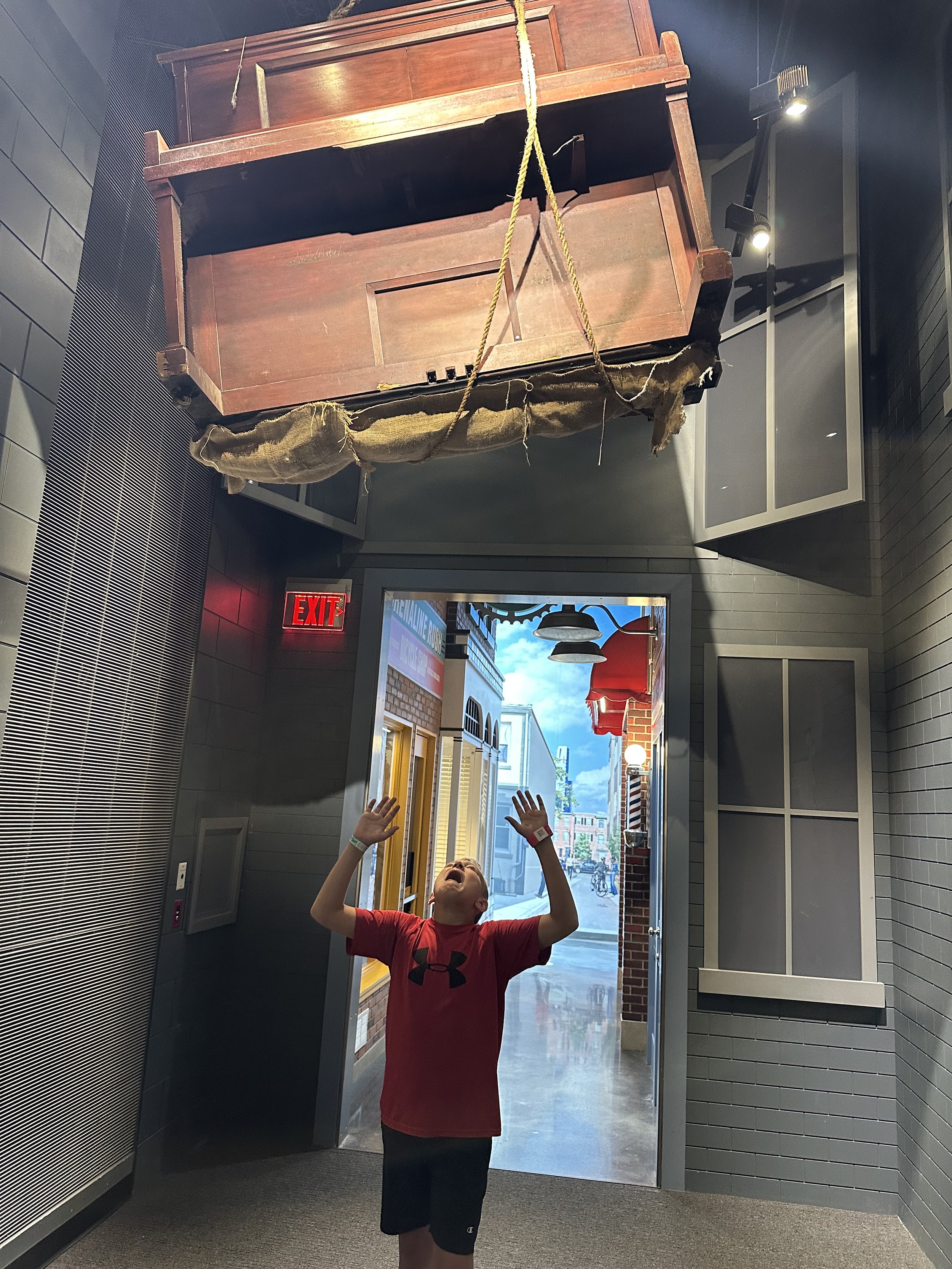 A young boy standing inside a building with his arms raised, looking up with excitement at a large, hanging wooden object resembling a coffin or chest, suspended by yellow ropes from the ceiling.