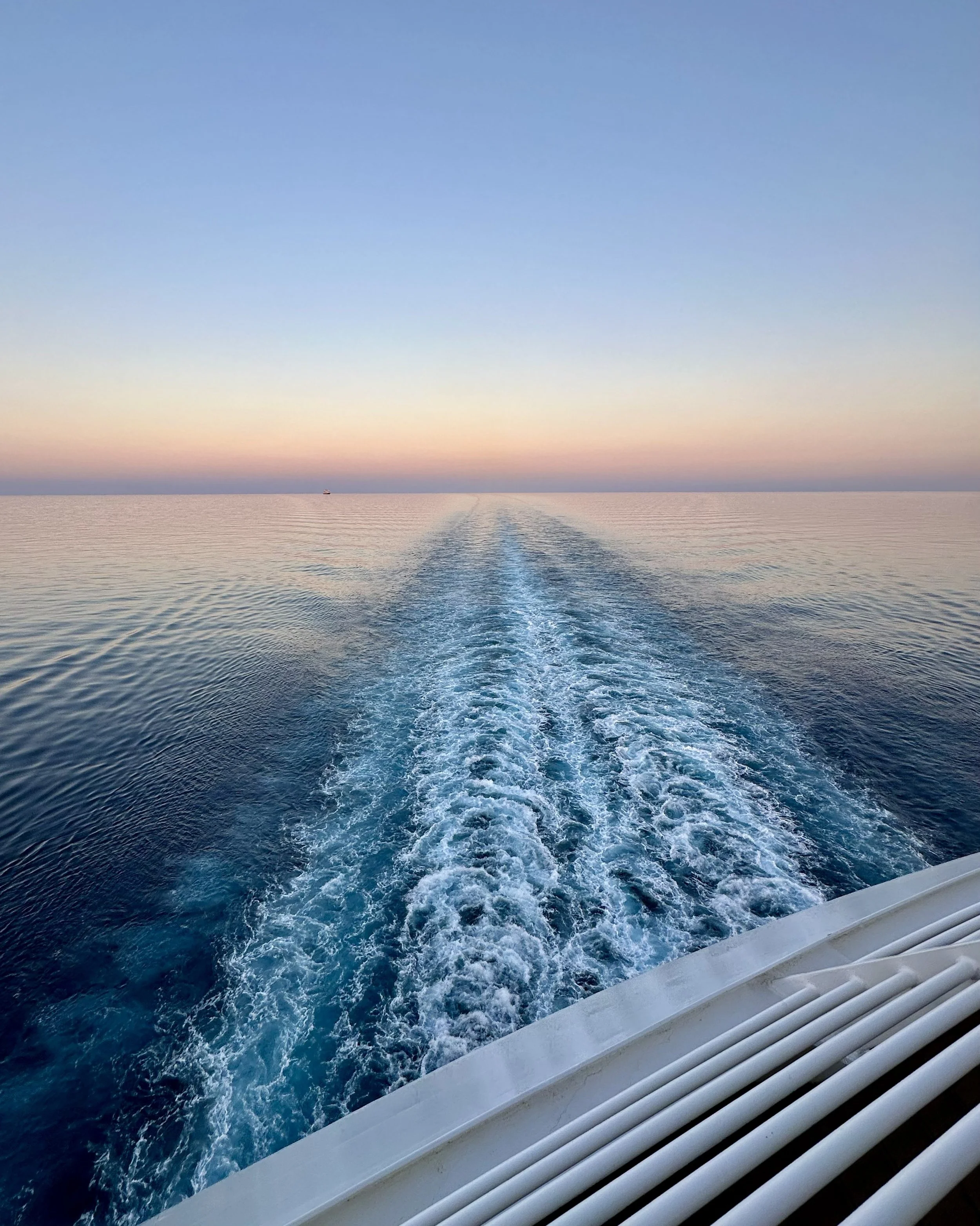View of the ocean from the back of a boat at sunset, with the boat's wake trail leading into the horizon and a small boat in the distance.