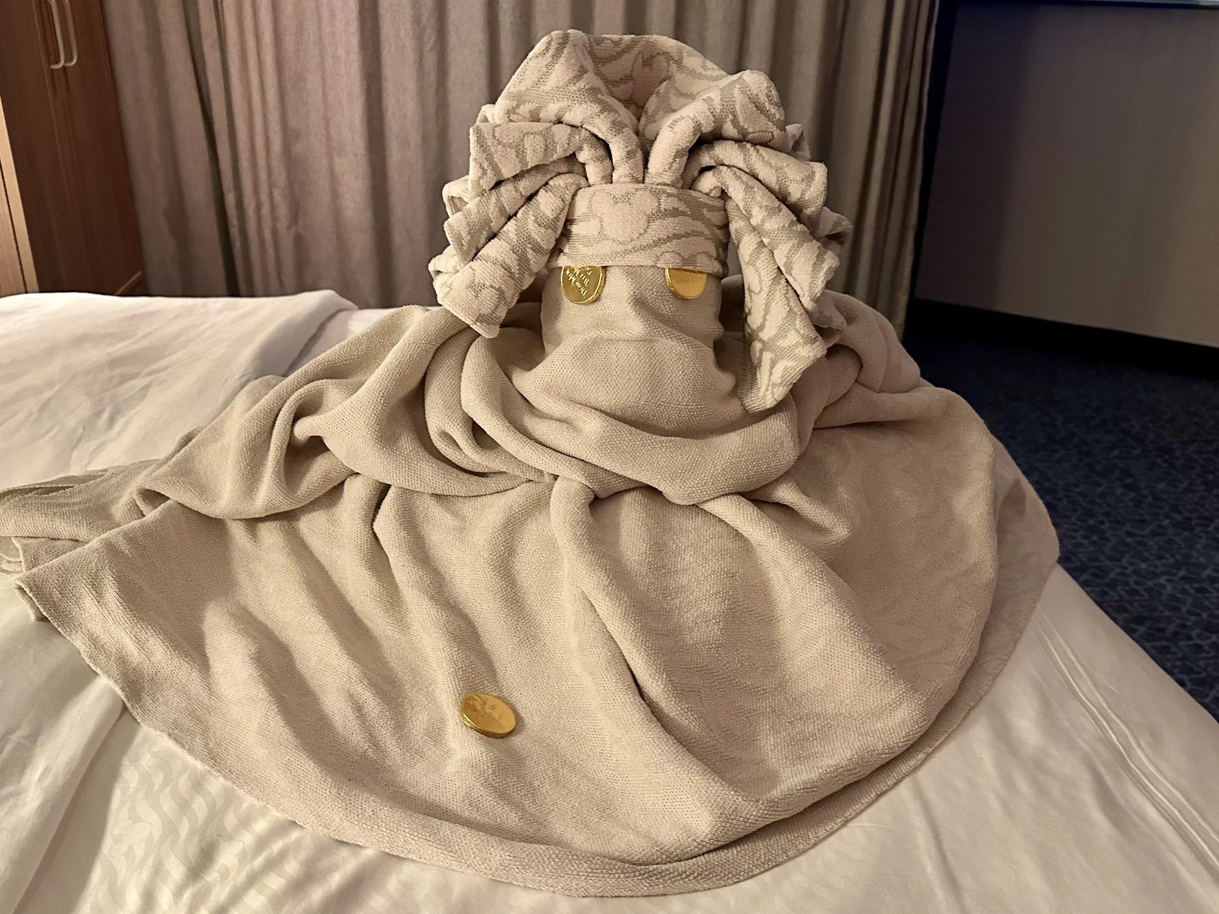 A towel art creation resembling a human figure, with a towel sculpted to have a head, arms, and body, placed on a bed with a beige bedspread. The sculpture has gold-colored coins as eyes. The background includes curtains and part of a wooden headboar
