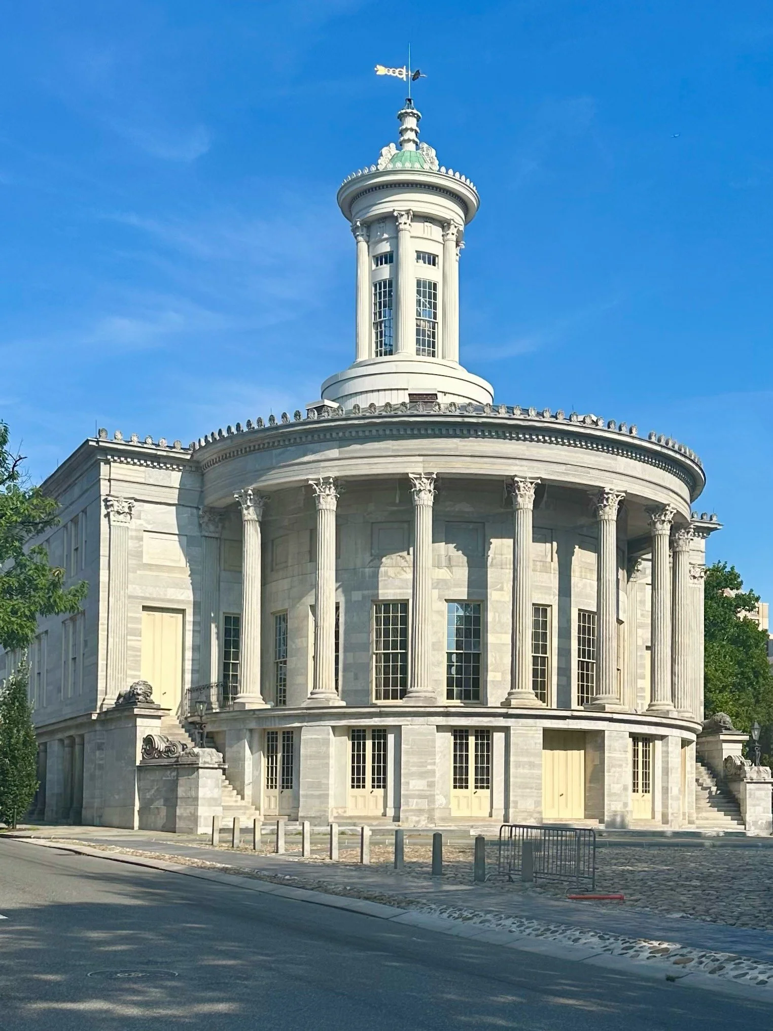 A historic white domed government building with tall columns, ornate details, and a weather vane on top, set against a clear blue sky.